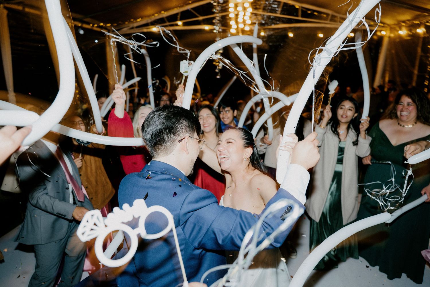 Guests dancing and celebrating at a lively wedding reception under decorative string lights and white fabric draping.