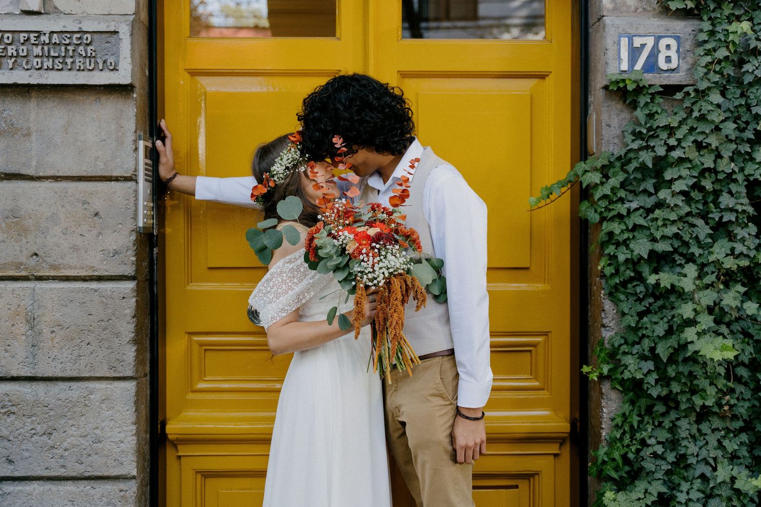 A couple shares a romantic moment in front of a bright yellow door with ivy growing on stone walls.