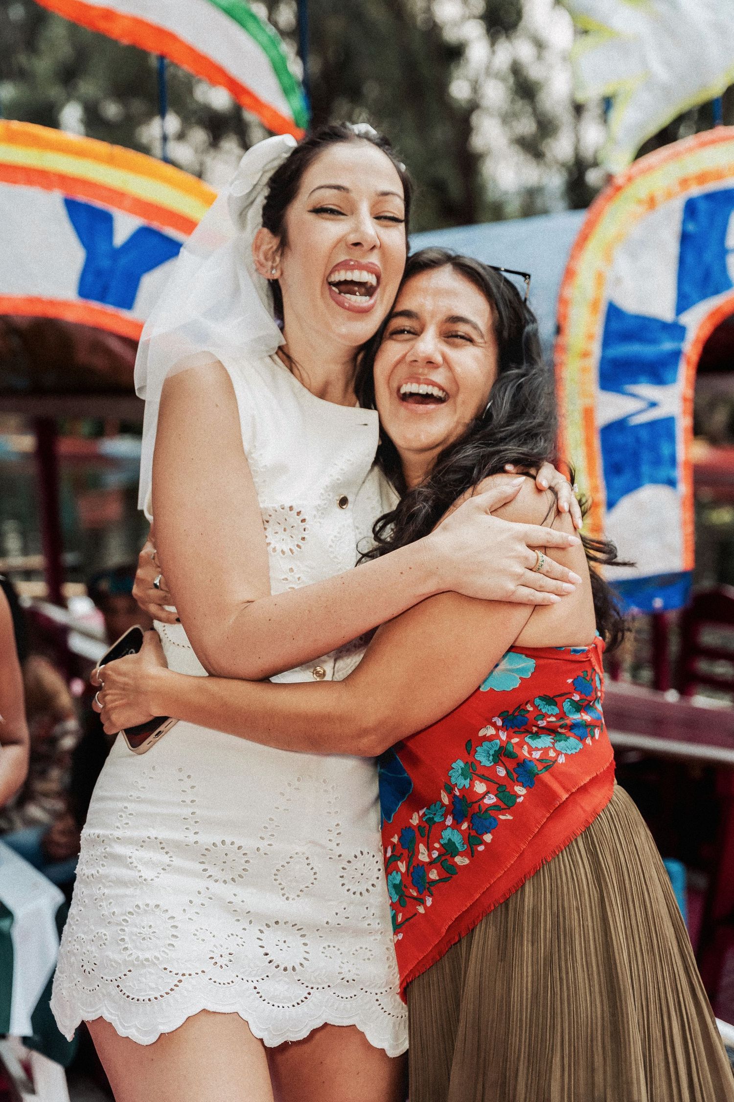 Two people share a joyful embrace and laugh together at an outdoor celebration with colorful decorations.