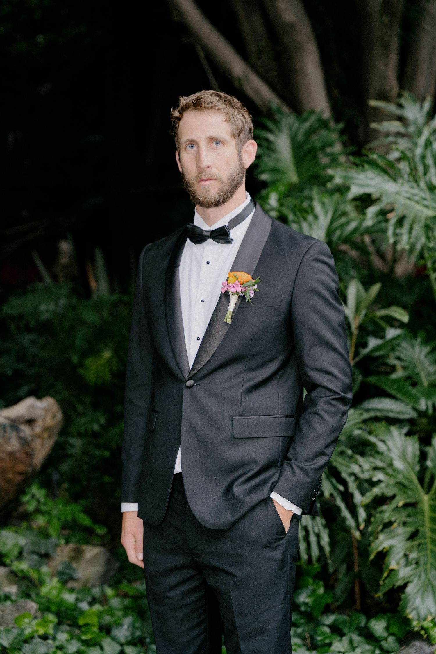 A groom in a classic black tuxedo and white bowtie poses against lush green foliage at a formal wedding.