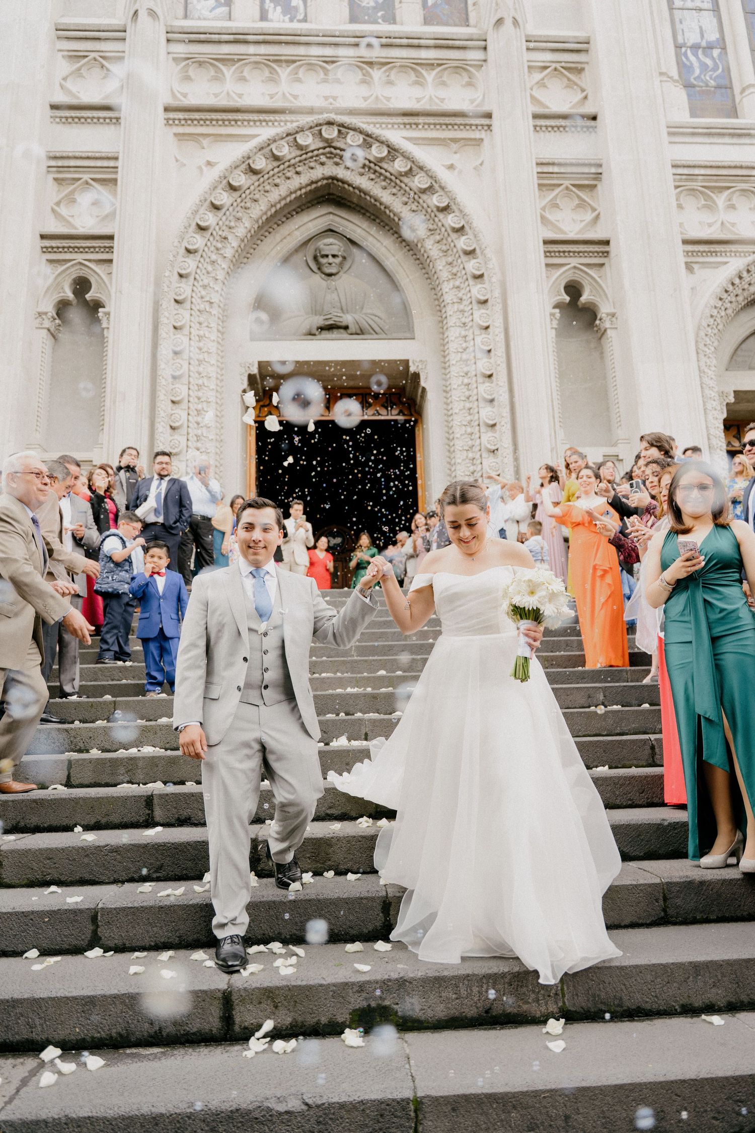 Wedding party celebrating on grand stone steps of ornate building as guests throw confetti during joyful exit.