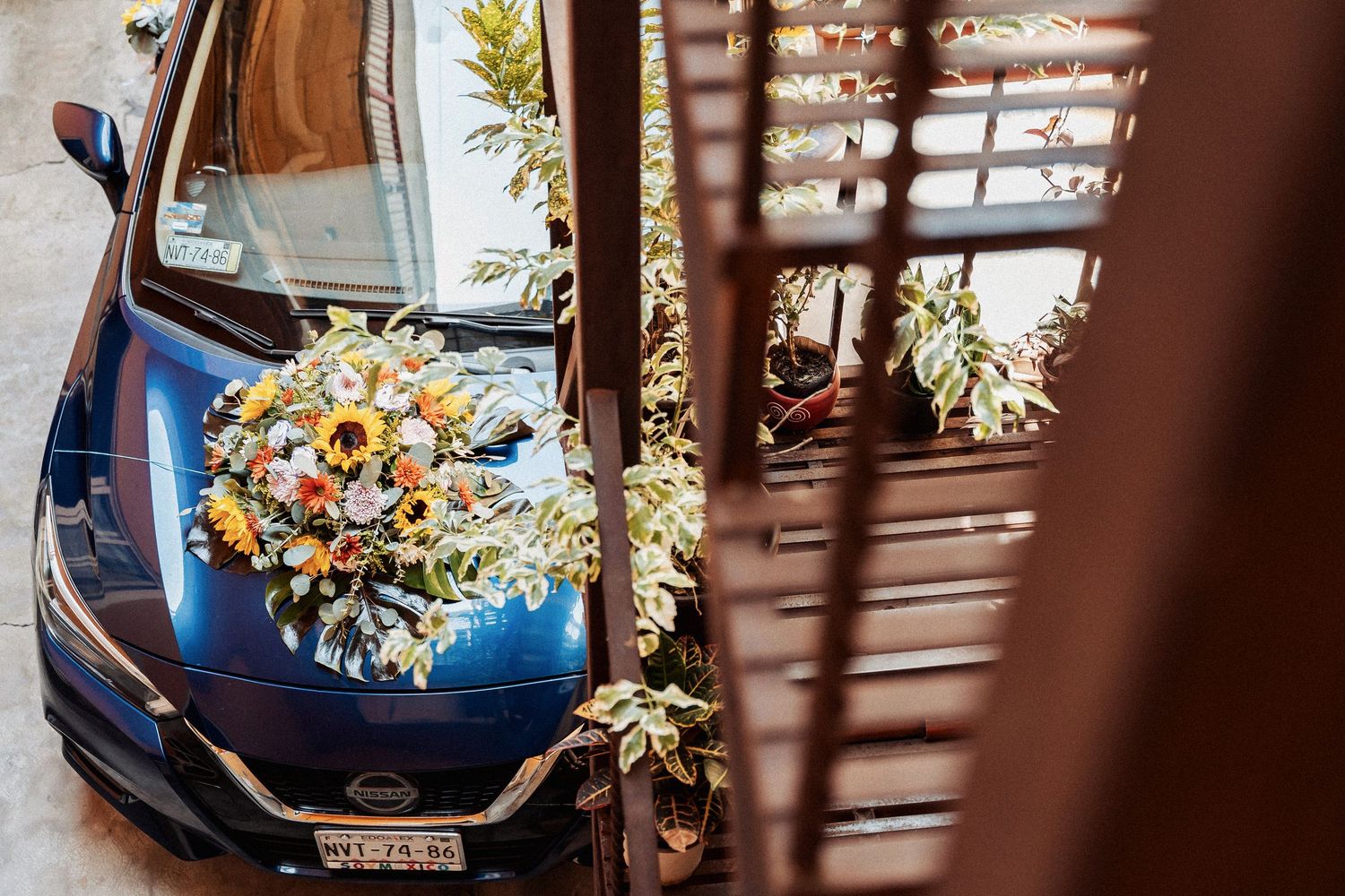 Wedding bouquet with white and yellow flowers laid on the hood of a blue car viewed through window blinds.