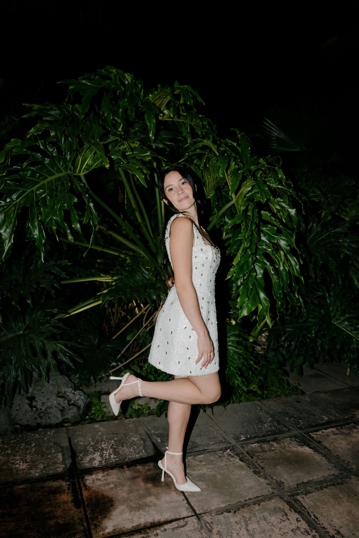 A person in a white dress and heels poses near lush greenery at night.