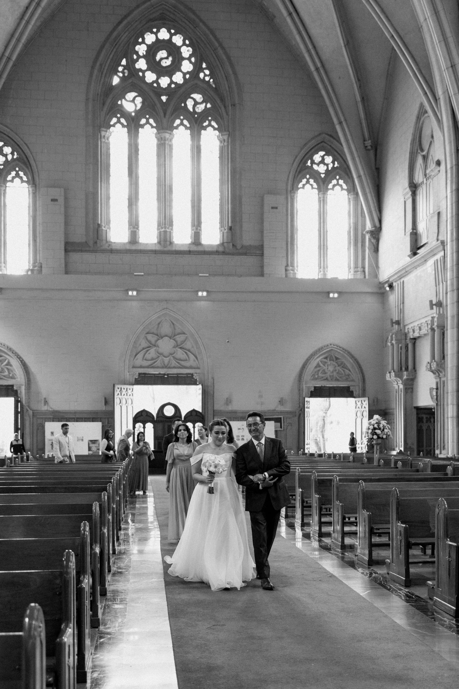 A wedding procession walks down the aisle of a Gothic church with vaulted ceilings and stained glass windows.