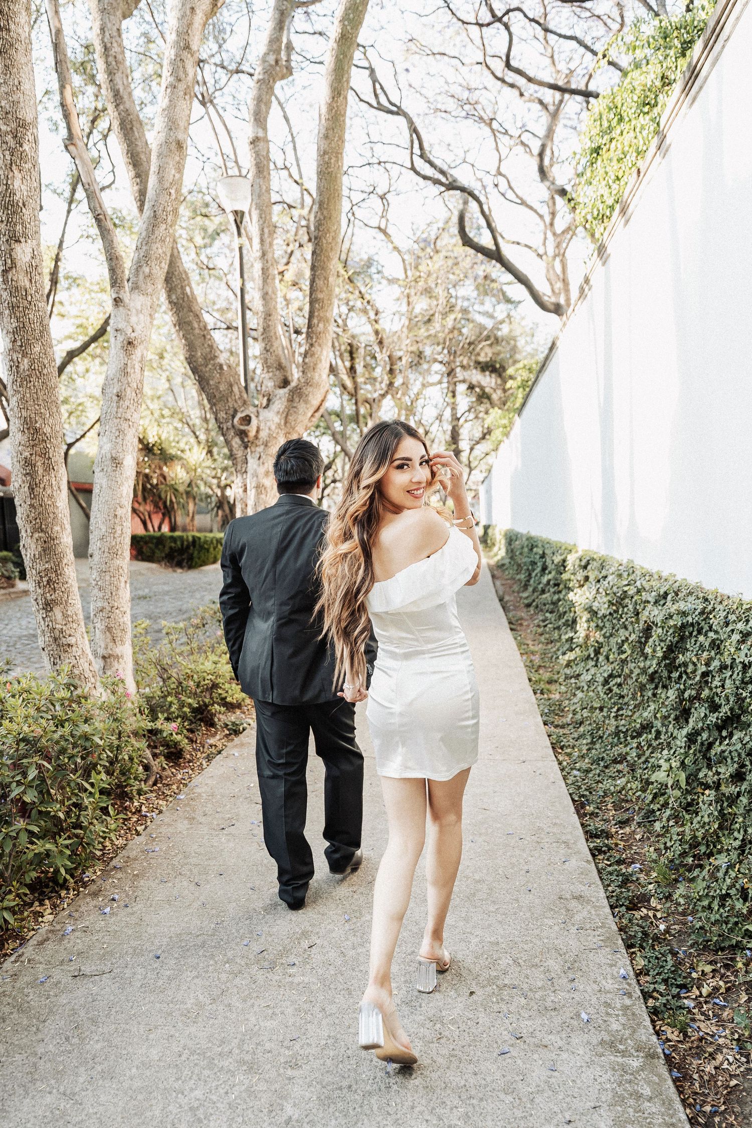 A couple walks along a garden path bordered by bare trees and white walls.