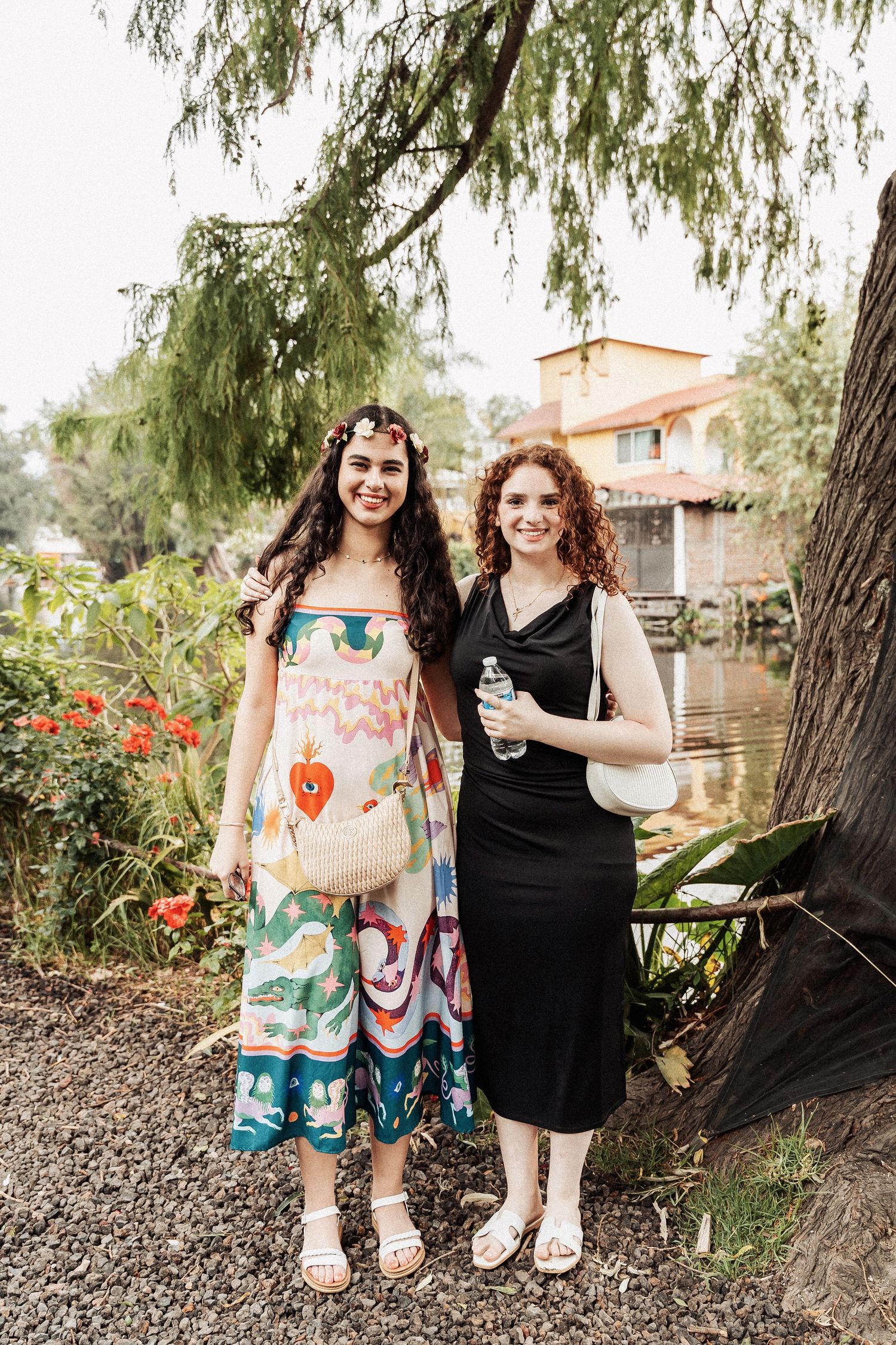 Two women smile together outdoors in contrasting dresses - one colorful and patterned, one solid black - near flowering plants.