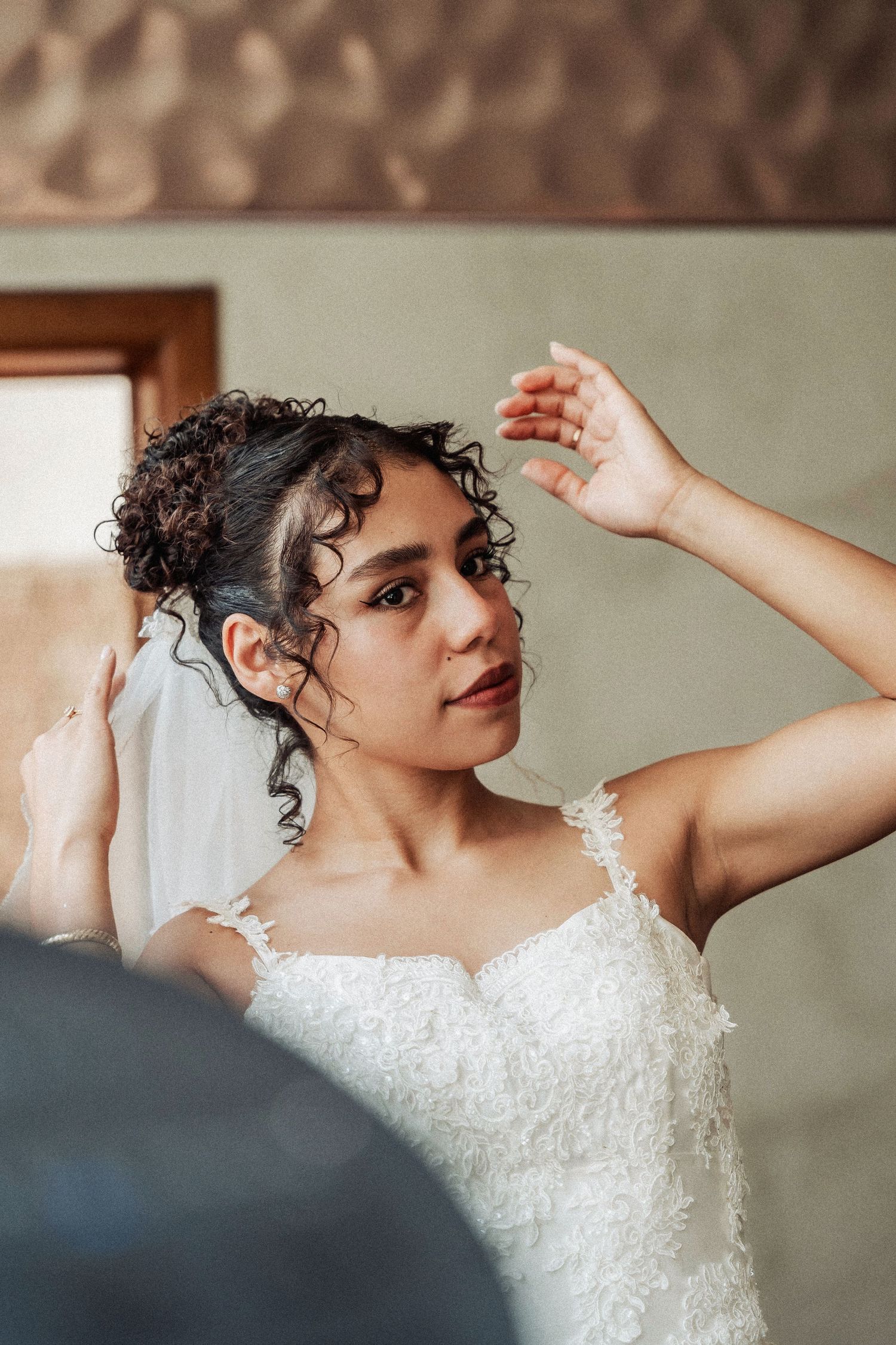 A person in a white lace wedding dress adjusts loose curls in their hair while wearing a veil.