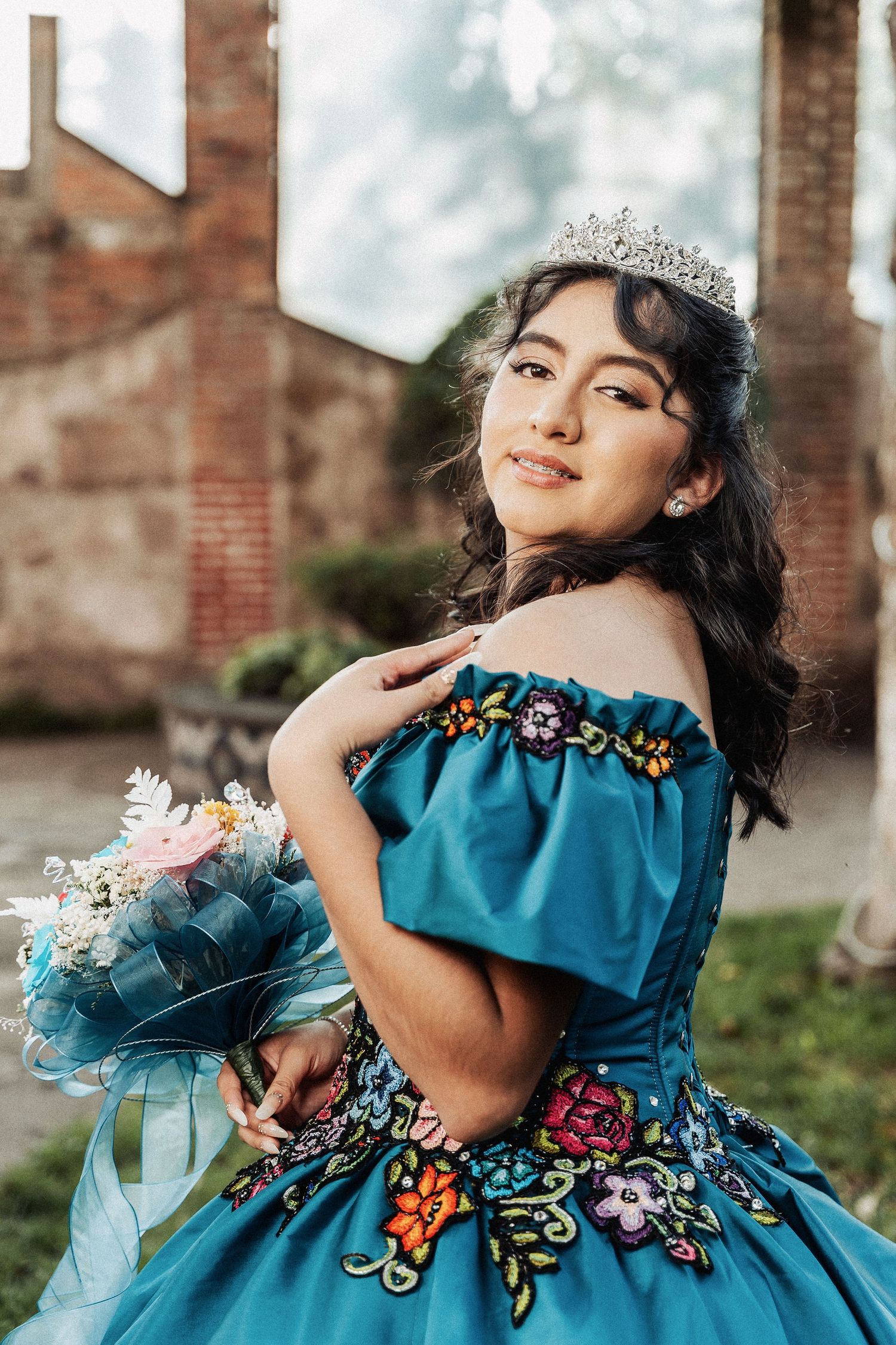 A quinceañera celebration featuring an elegant teal ball gown with floral embroidery, tiara, and matching bouquet.