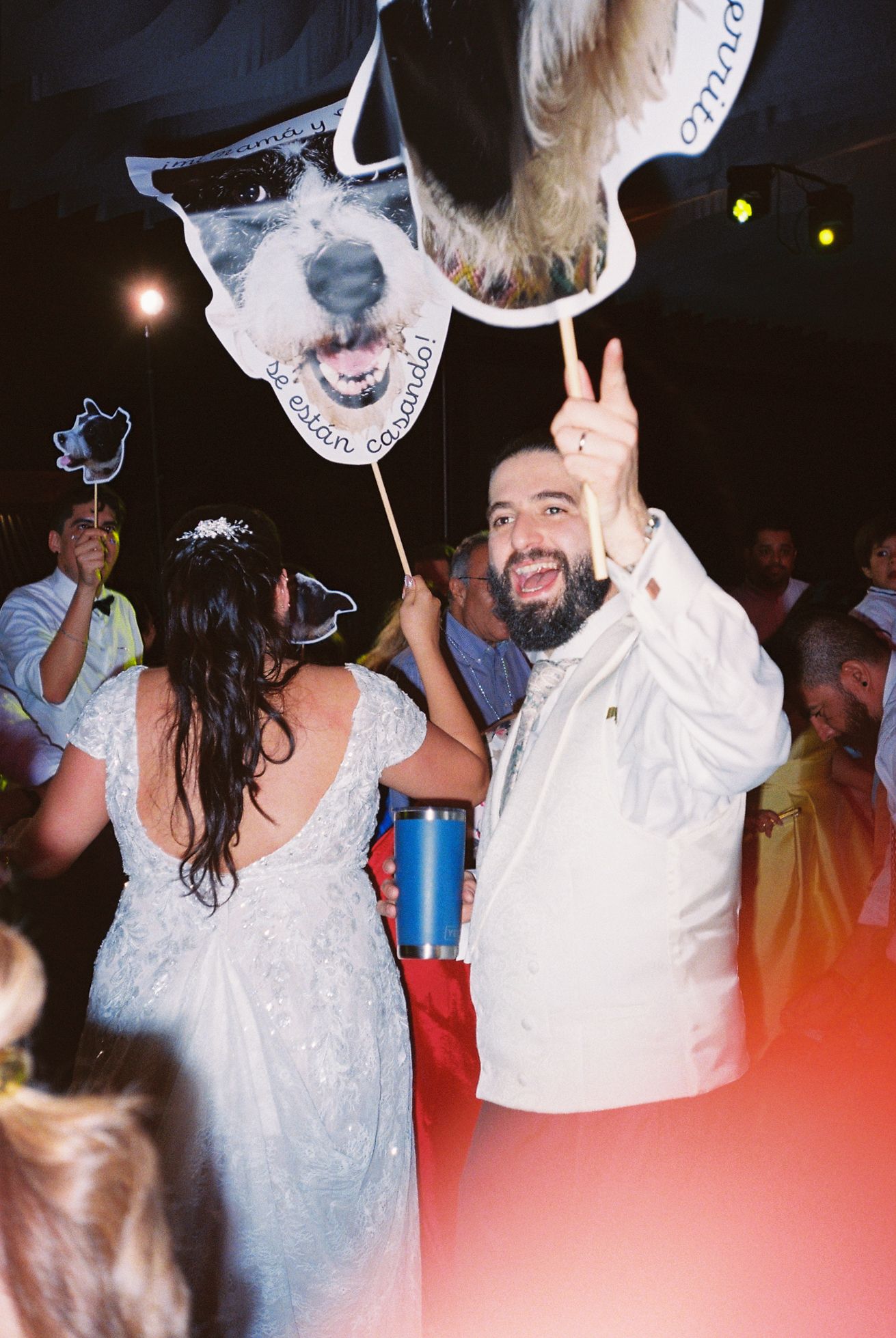 A couple celebrates at a party with a large dog-themed balloon and drinks while making a peace sign gesture.
