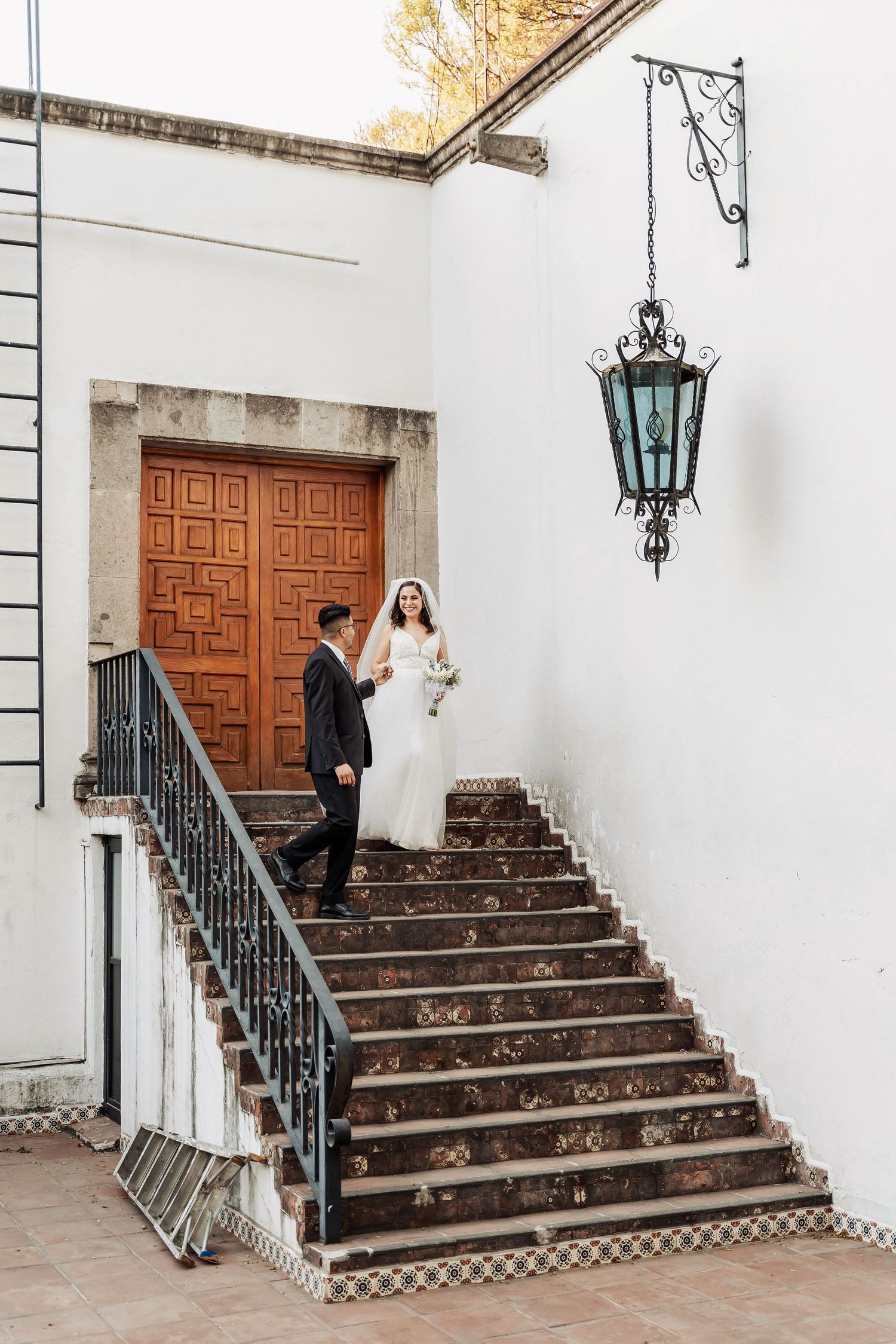 A couple stands on an elegant staircase with a wooden door and vintage lantern against a white Spanish colonial wall.