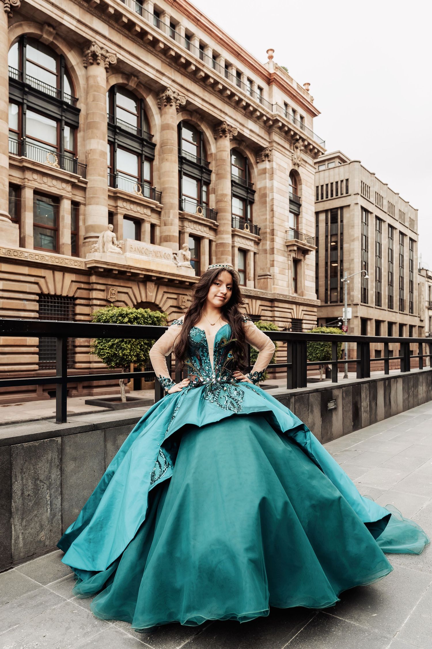 Woman in a dramatic turquoise ball gown poses elegantly against a classical stone building exterior.