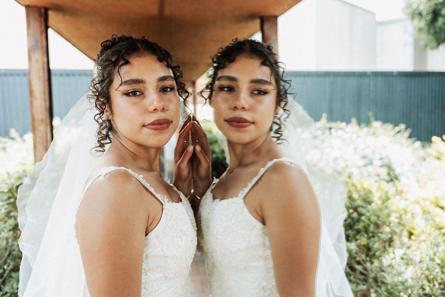 Two curly-haired young women in matching white spaghetti strap dresses stand together outdoors in natural lighting.