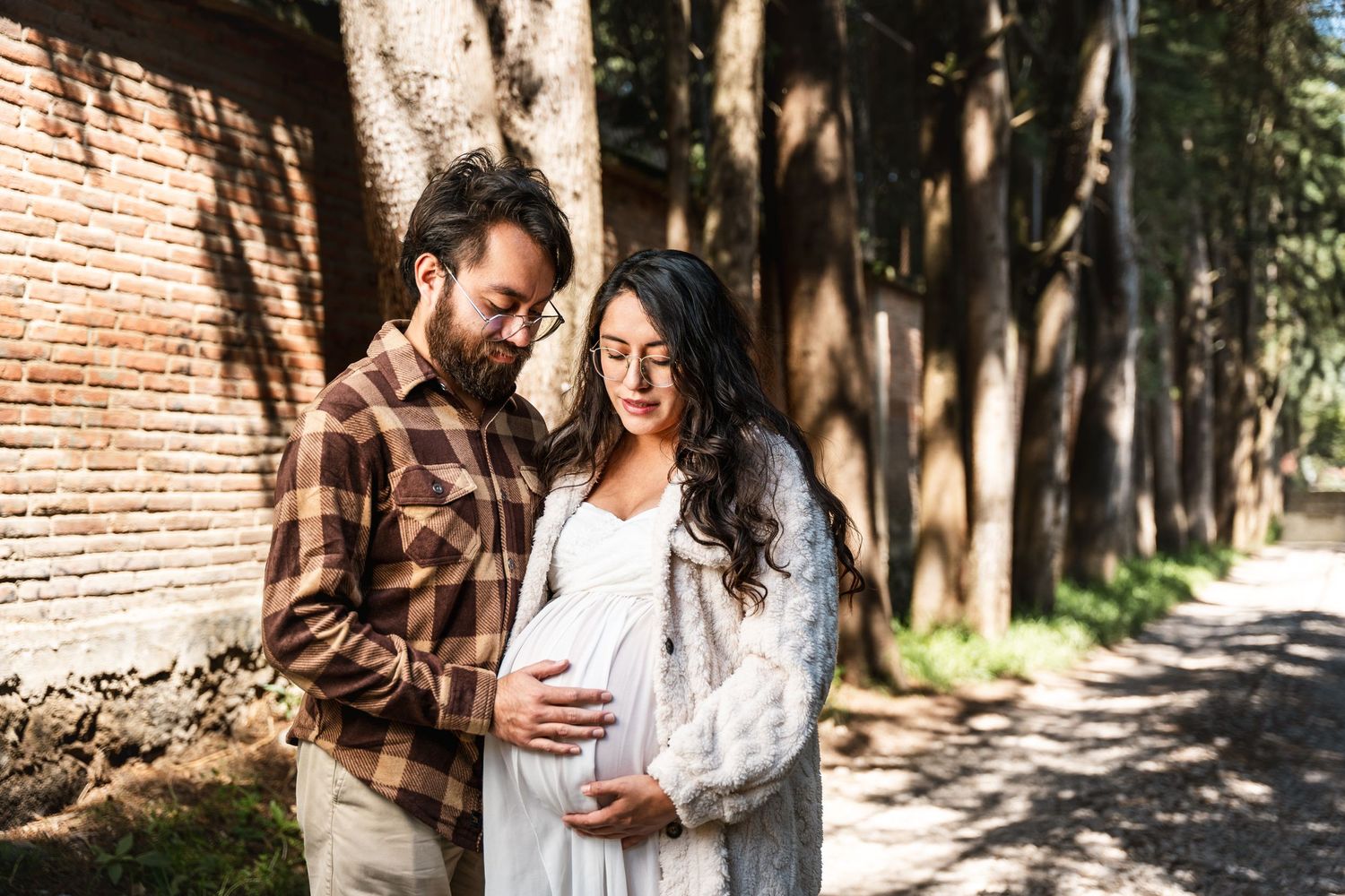 A maternity photo of a couple standing in dappled sunlight against a brick wall.