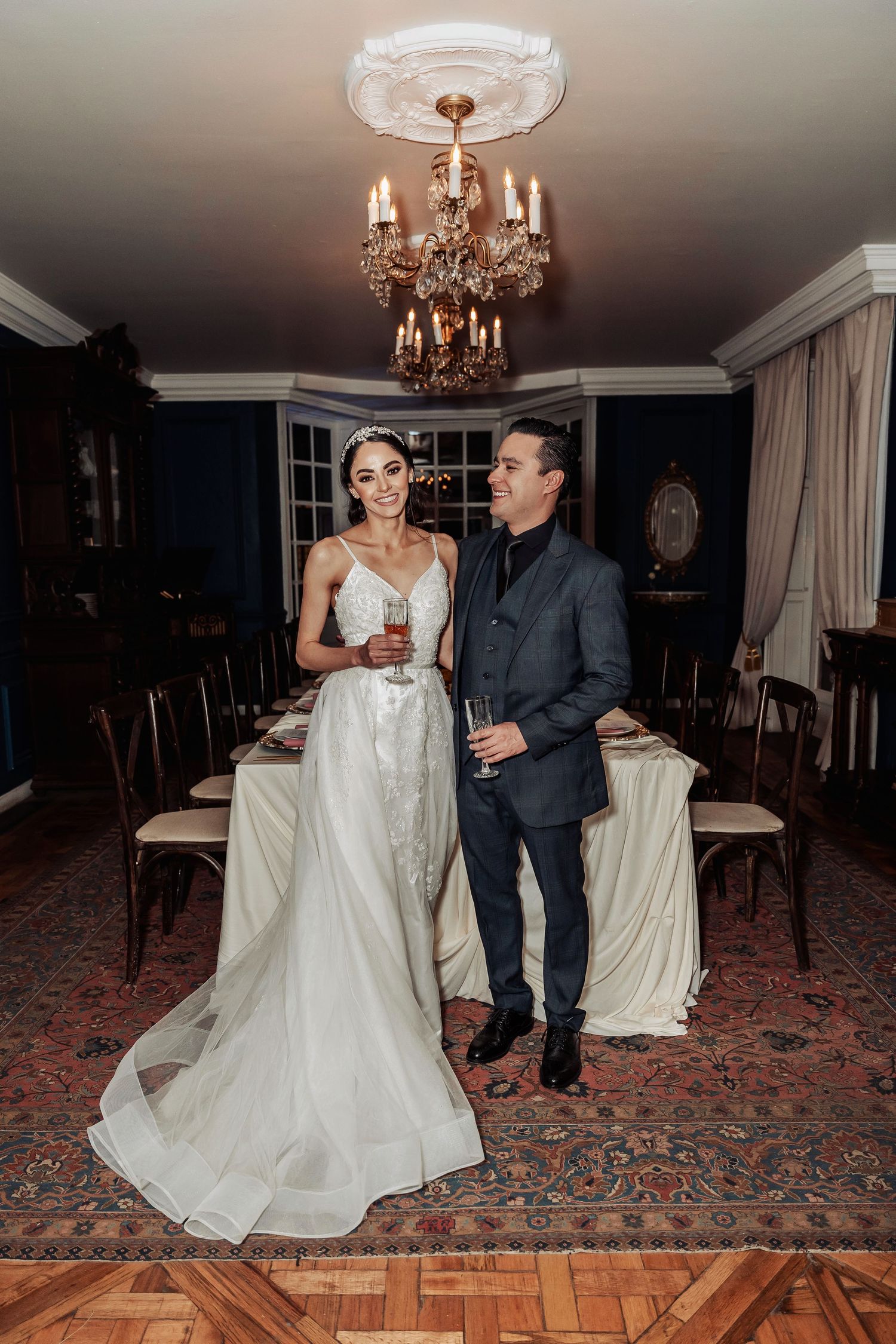 A couple in formal attire stands in an elegant dining room under a crystal chandelier.
