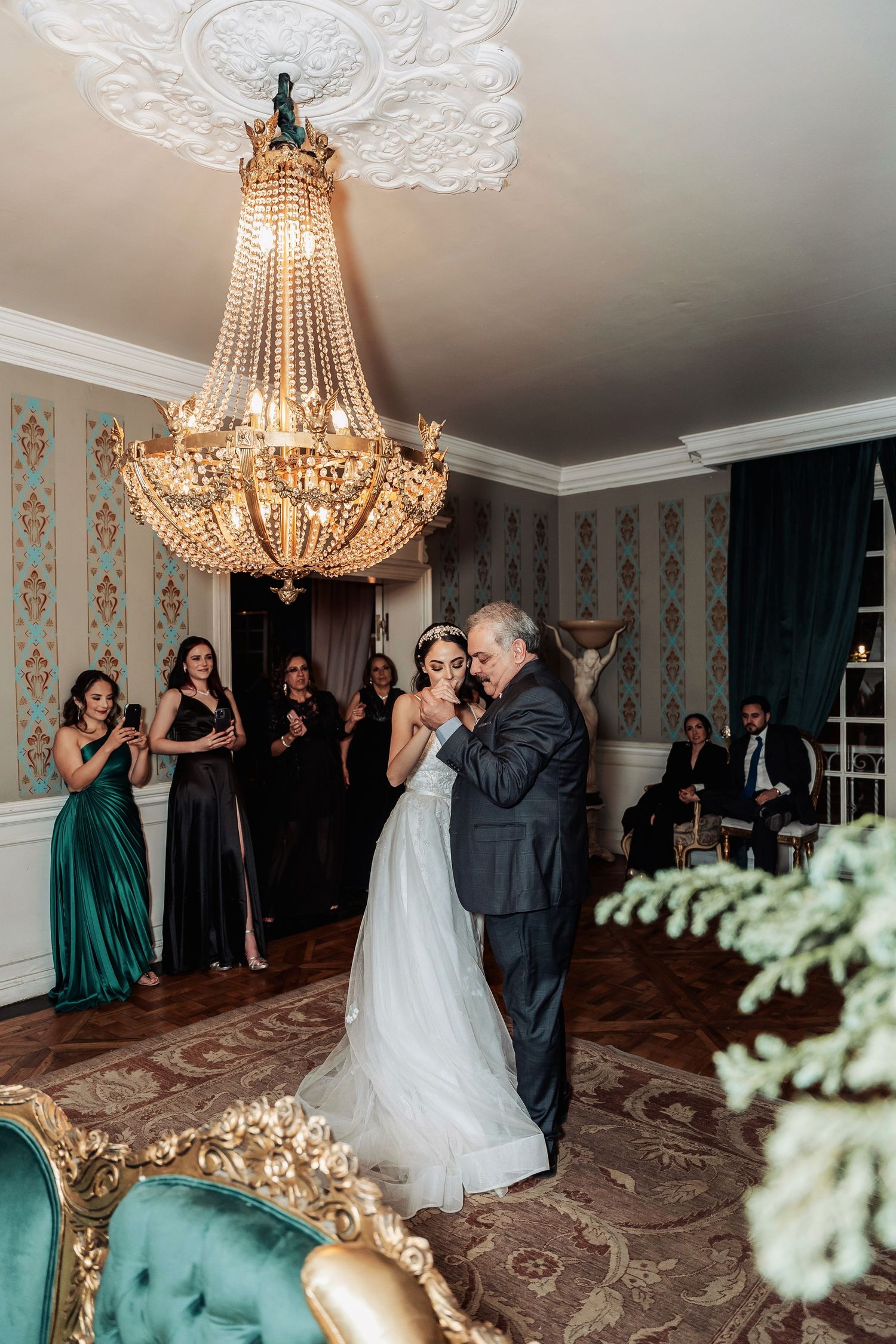 A romantic first dance under an elegant crystal chandelier in an ornate room with vintage wallpaper and green decor.