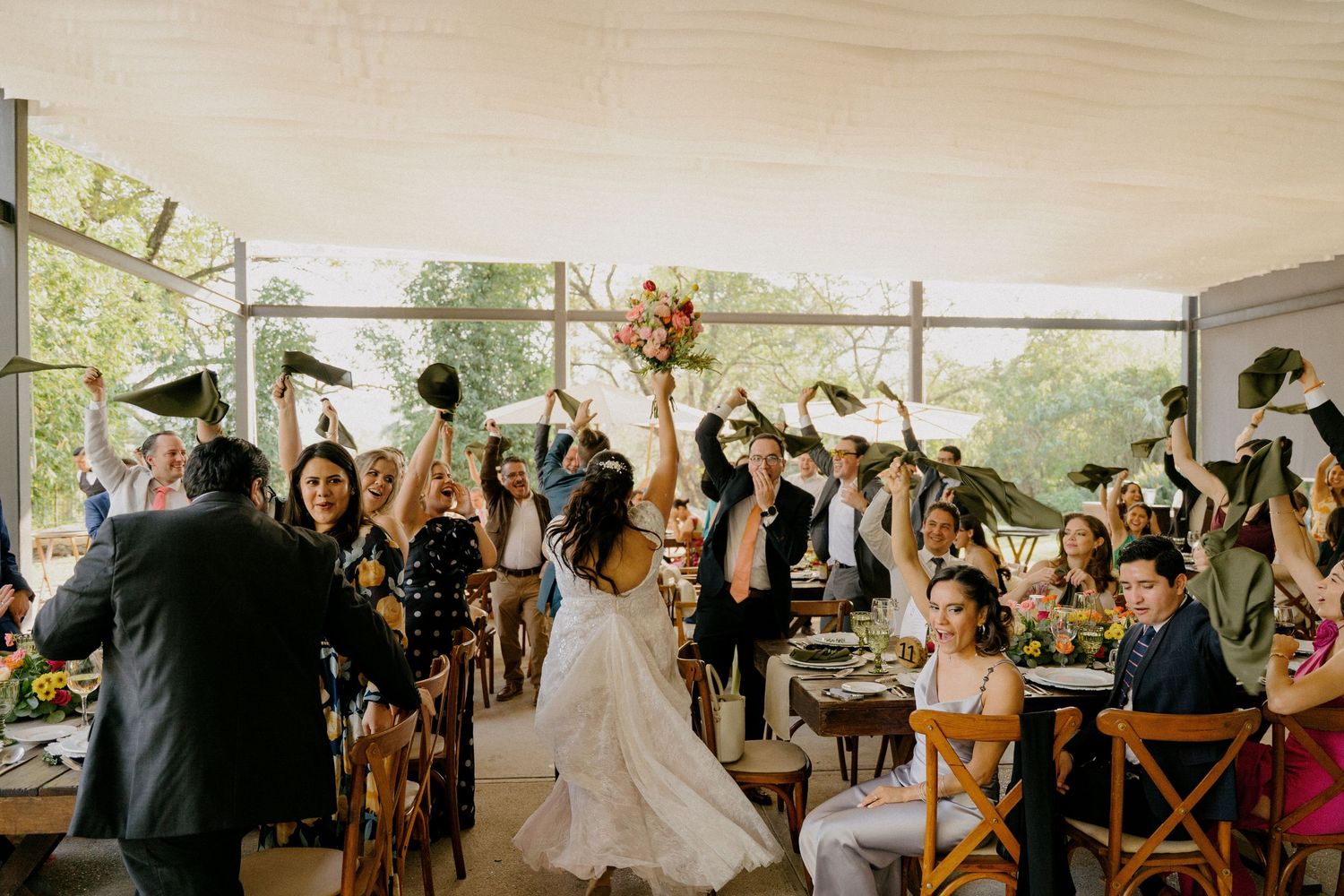 Wedding guests cheer and celebrate with raised arms during a reception in a bright sunlit venue.