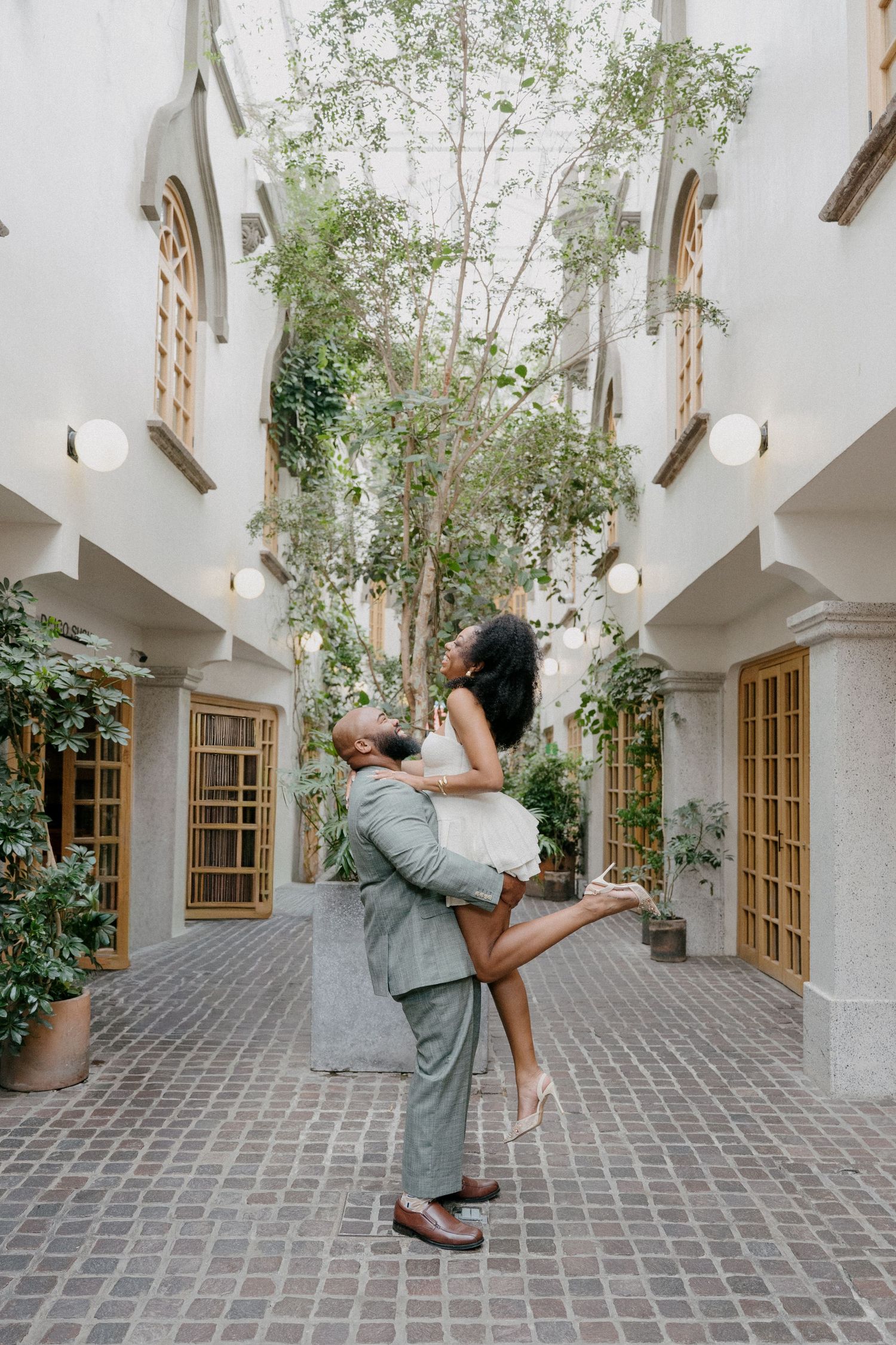 A romantic couple in matching green outfits shares a tender moment in a courtyard with stone walls and wooden windows.