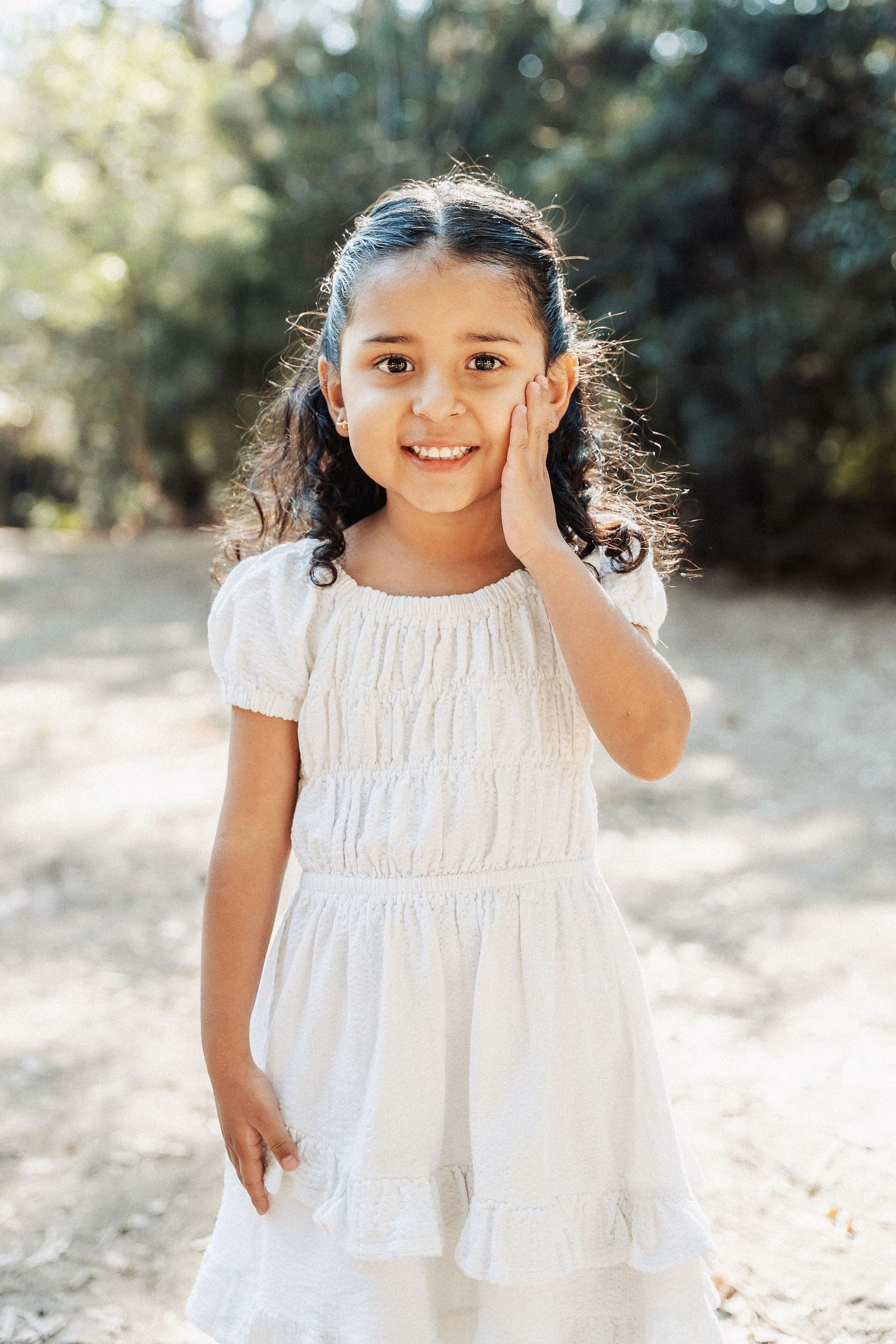 Young child in a white summer dress stands outdoors with hand on cheek in natural lighting.