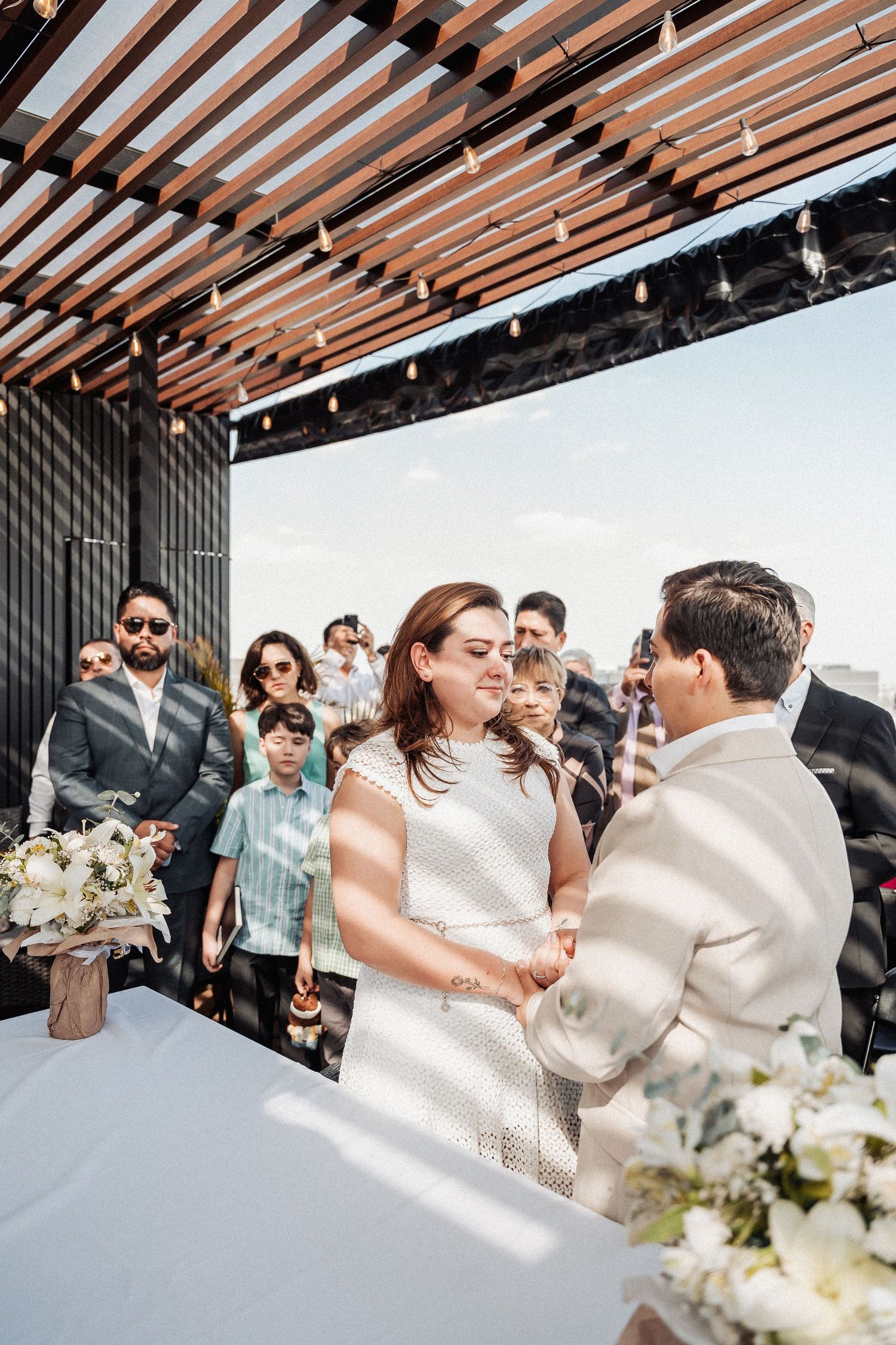 A wedding ceremony under a wooden pergola with guests watching the exchange of vows.