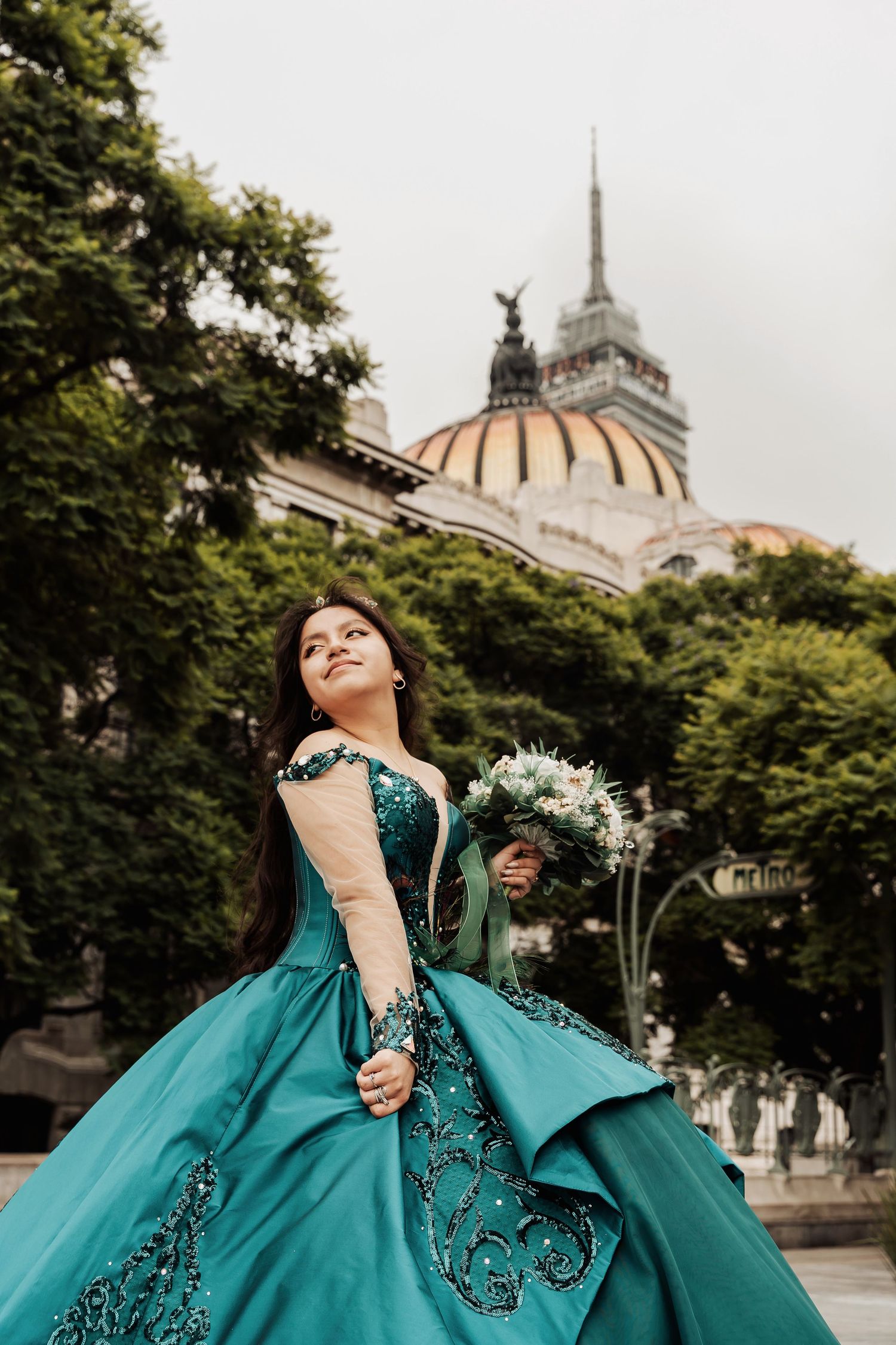 A person in an elegant teal quinceañera dress with black embroidery poses in front of a historic domed building.