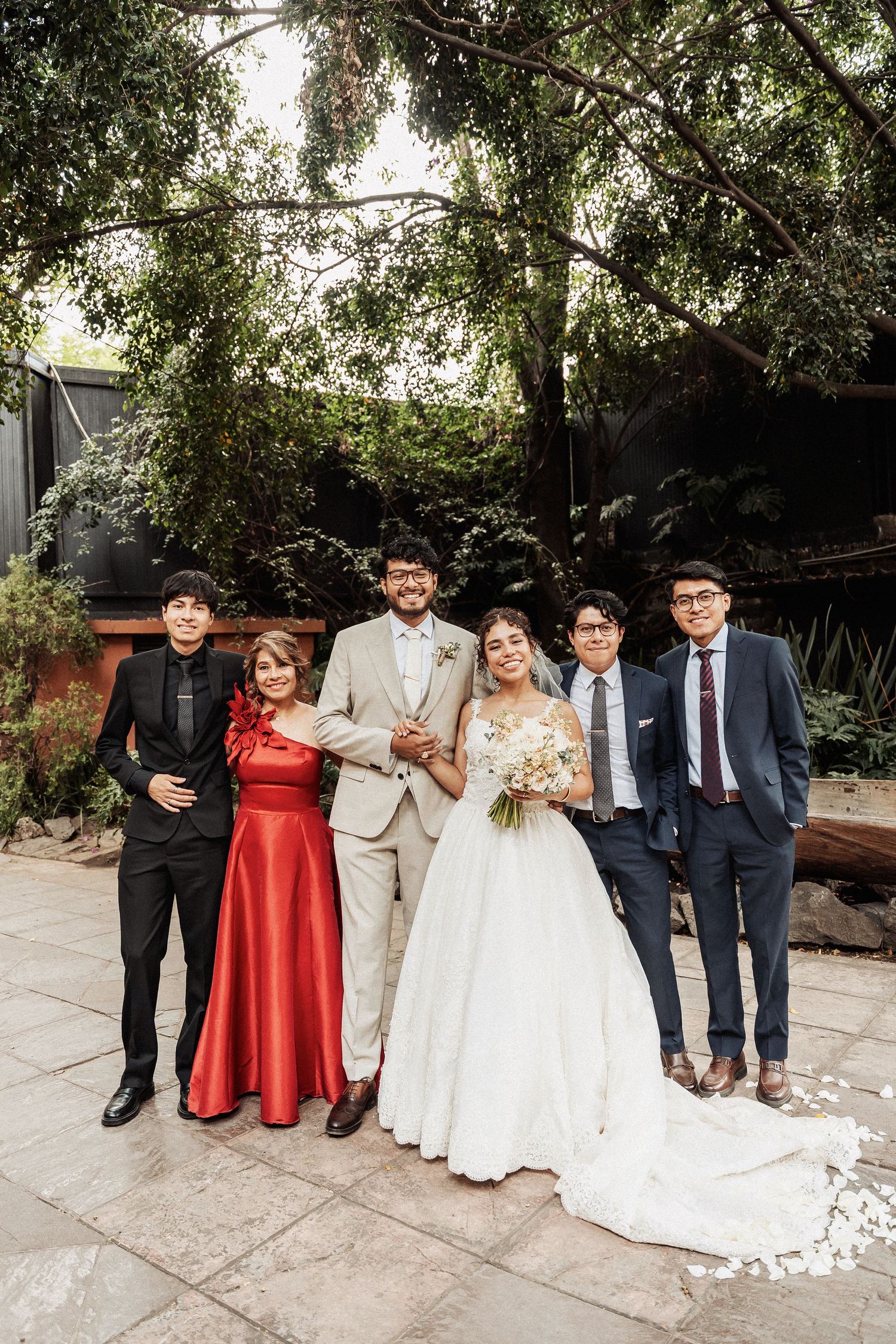 Wedding party poses outdoors under trees with couple in white and beige and guests in formal attire.