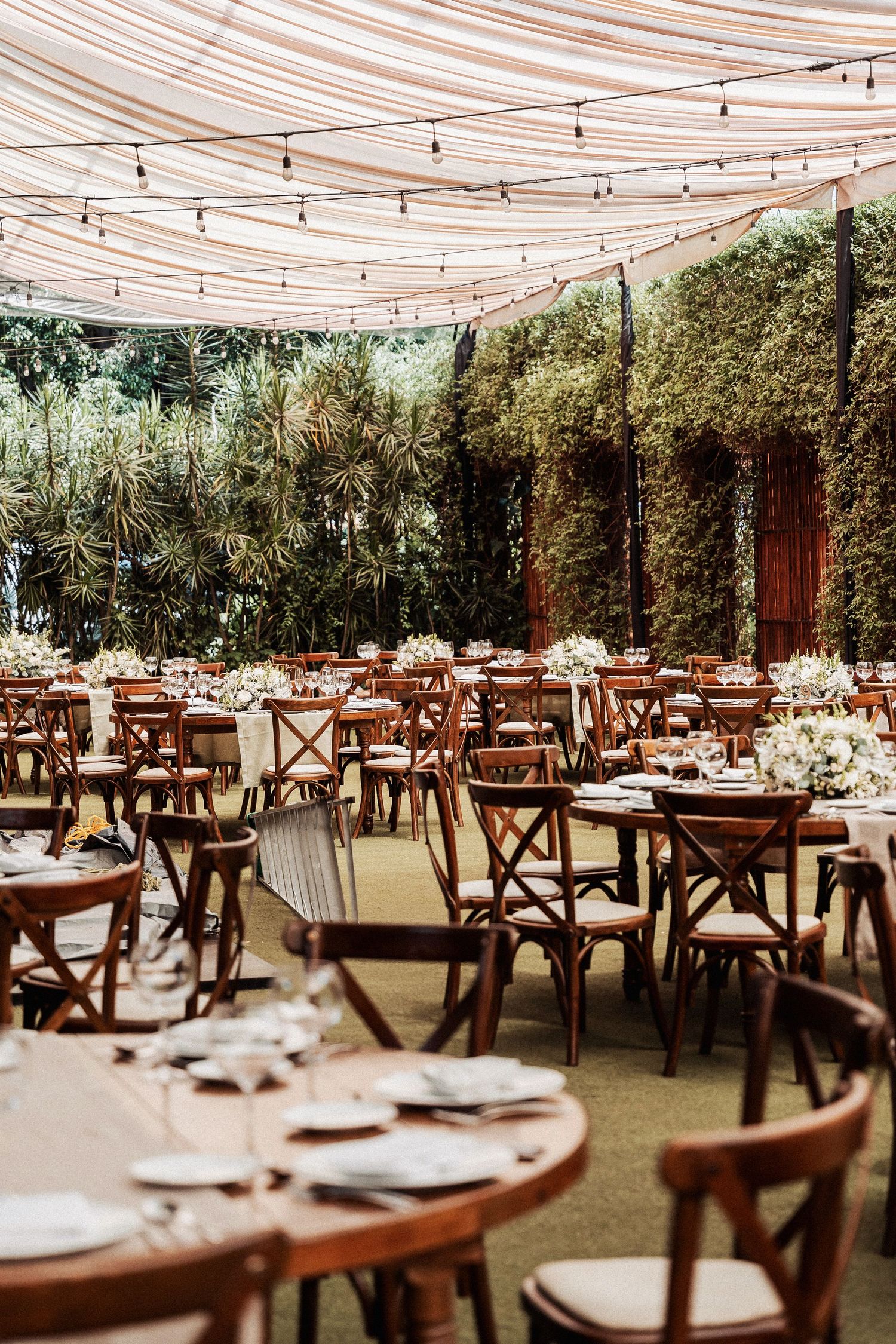 Outdoor wedding reception venue with wooden chairs, round tables, and draped fabric ceiling with string lights.