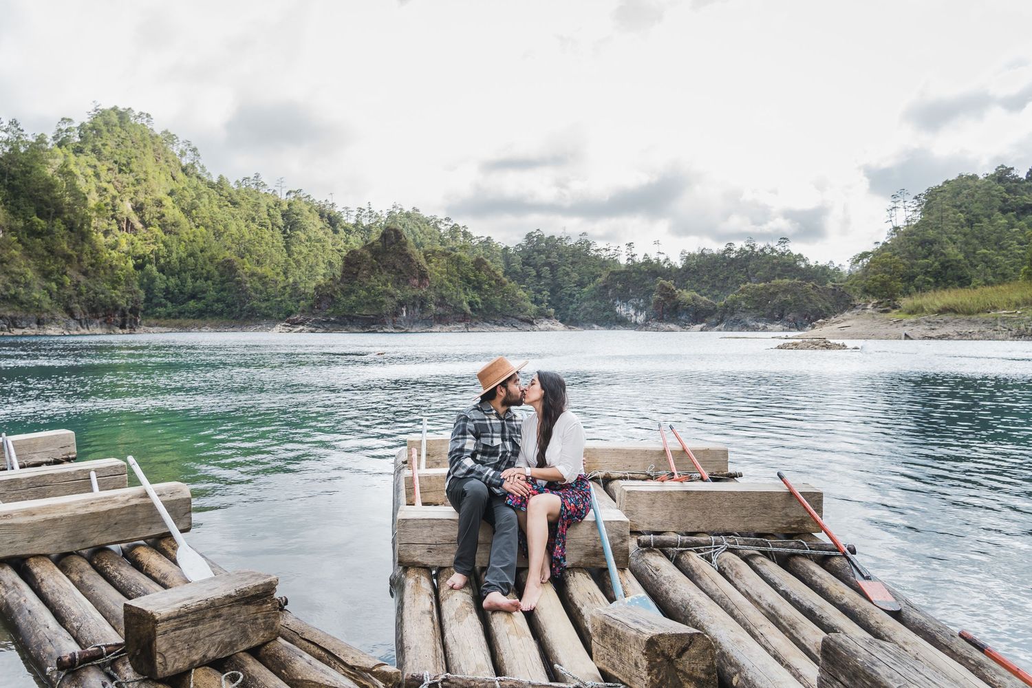 A couple sits on a wooden dock overlooking tranquil waters surrounded by lush greenery.