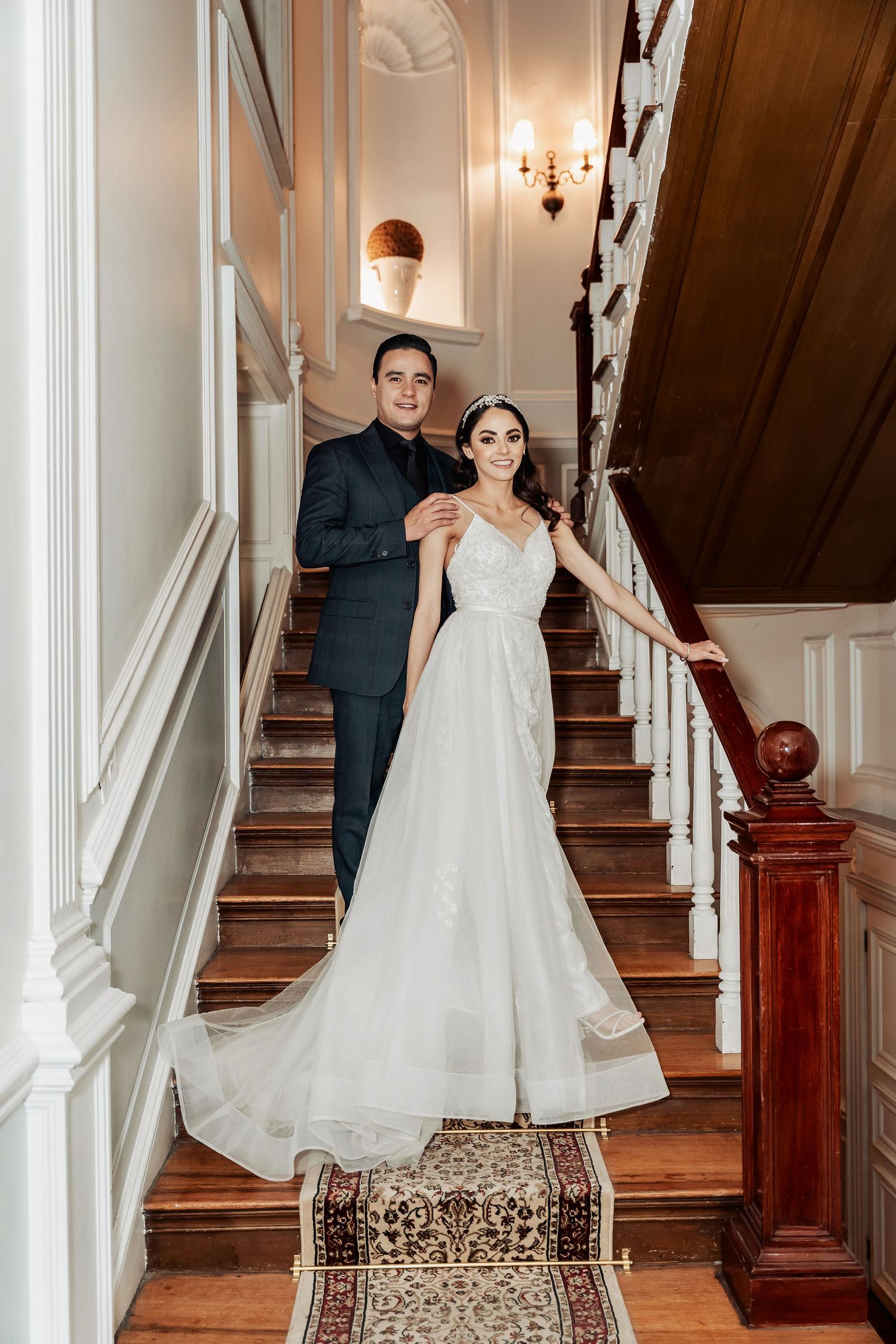 A couple poses on an elegant wooden staircase in classic wedding attire.