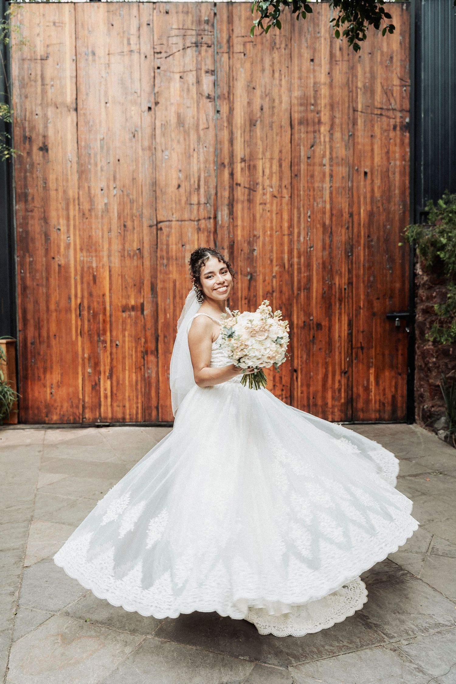 A bride in a flowing lace wedding gown twirls to show off her dress against a rustic wooden door backdrop.