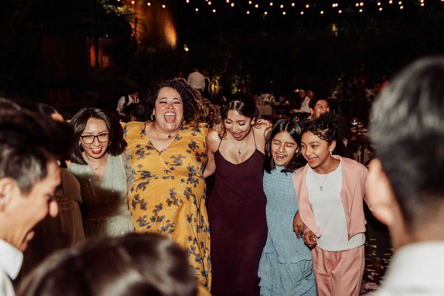Festive wedding party scene with guests enjoying themselves under string lights at an indoor venue.