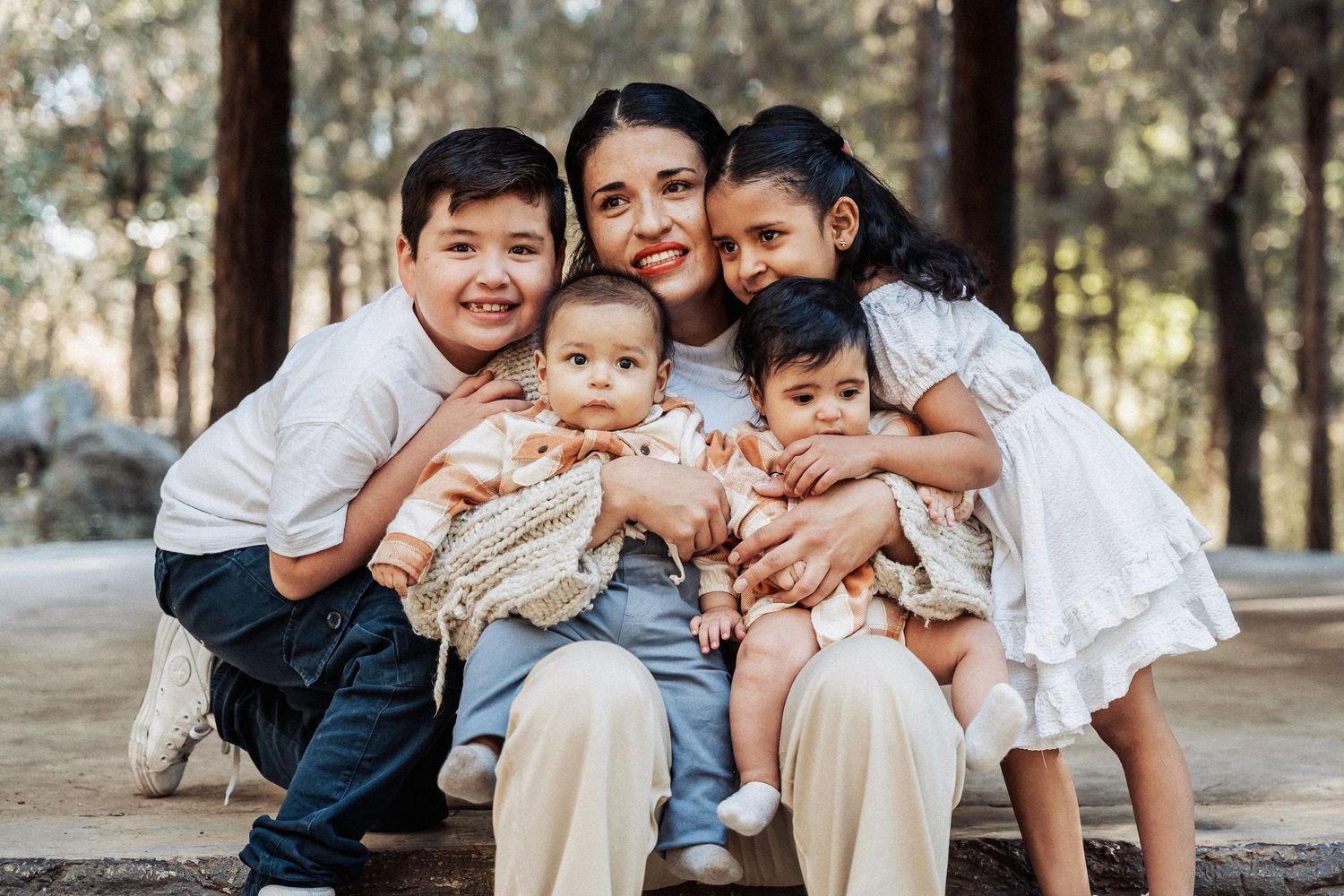 A family of five sits close together outdoors in a forest setting on a sunny day.