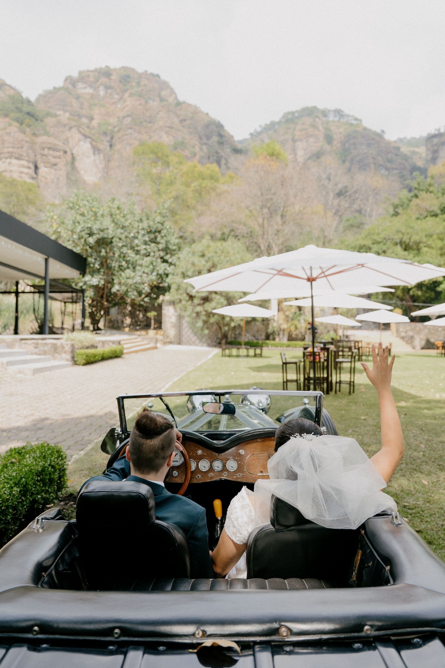 Two people riding in a vintage convertible car at an outdoor venue with white umbrellas and mountain backdrop.