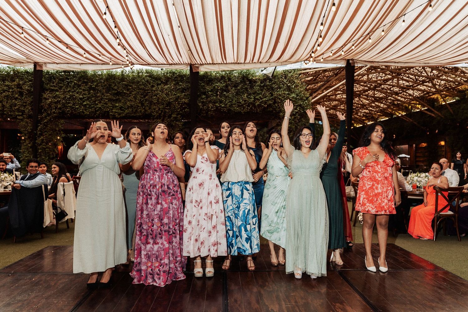 Wedding guests dancing in colorful dresses under a draped tent ceiling at an evening reception.