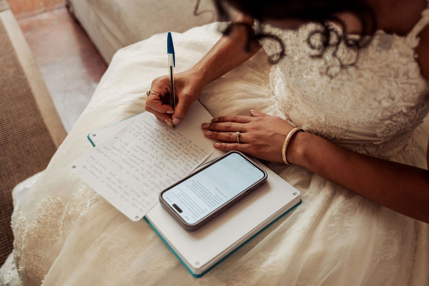 Person writing in a journal with a smartphone and notebook on a white surface.