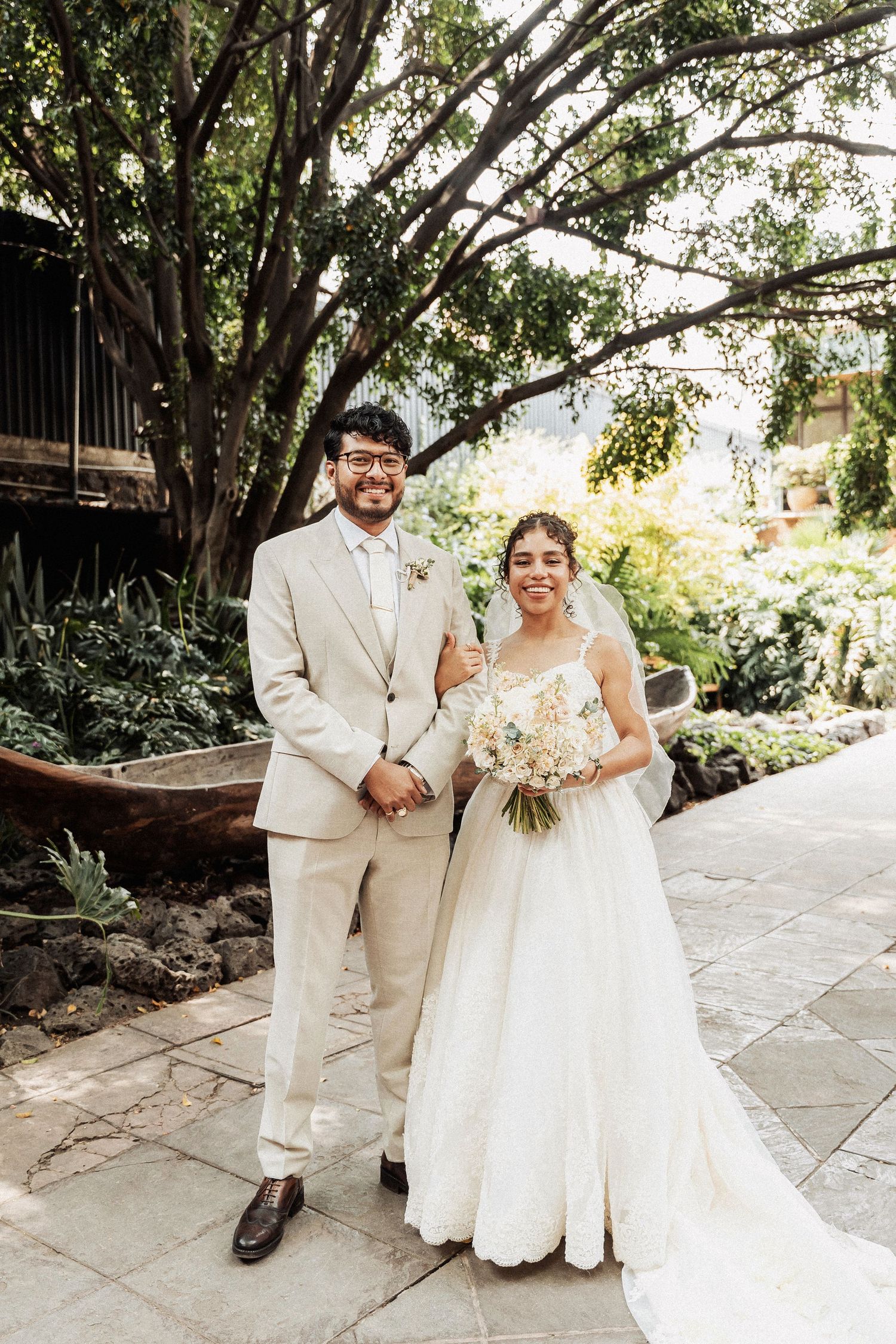 A joyful couple on their wedding day stands together outdoors, with the groom in beige and bride in white holding a bouquet.