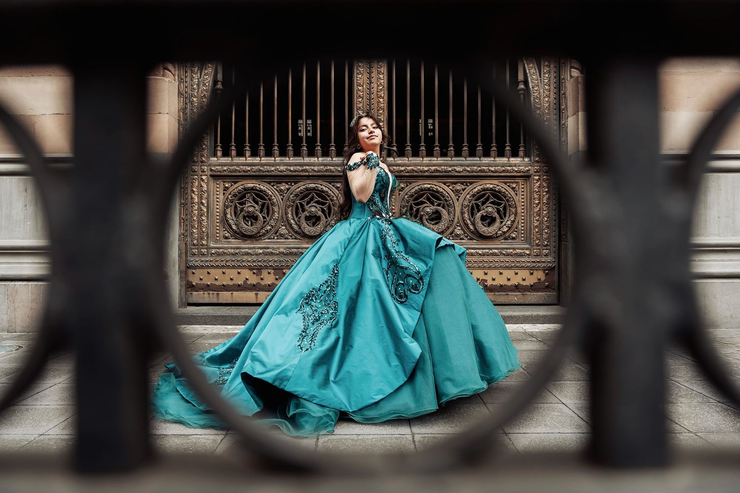 A figure in a stunning turquoise ball gown poses elegantly against an ornate architectural backdrop with decorative gates.