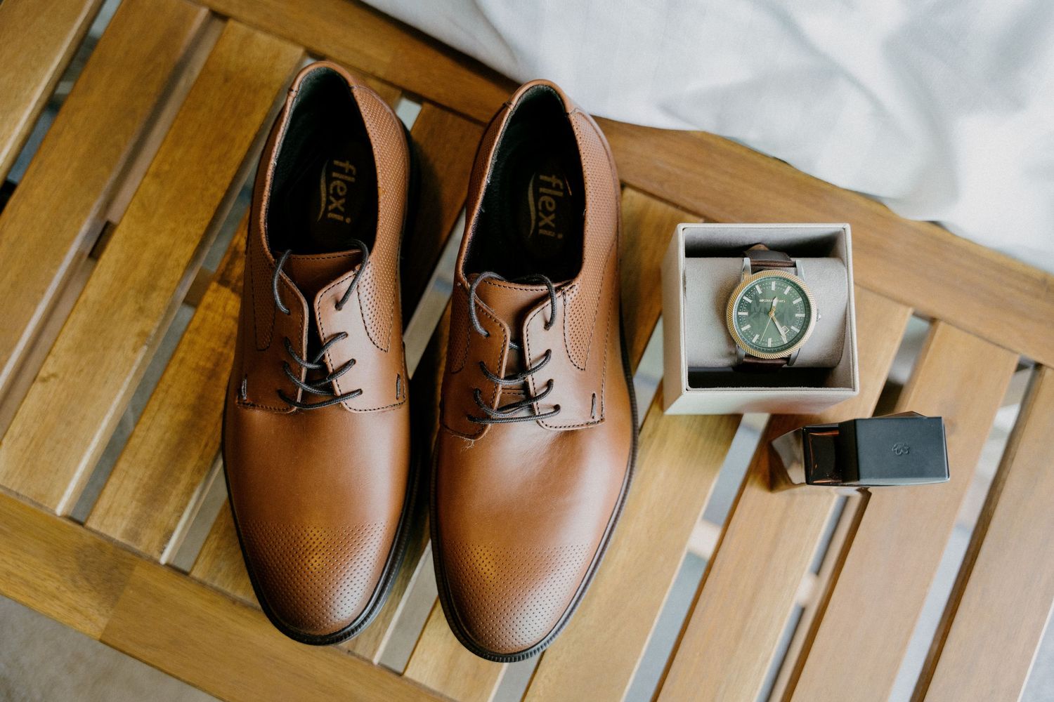 Brown leather dress shoes and a wristwatch displayed on a wooden slatted surface.