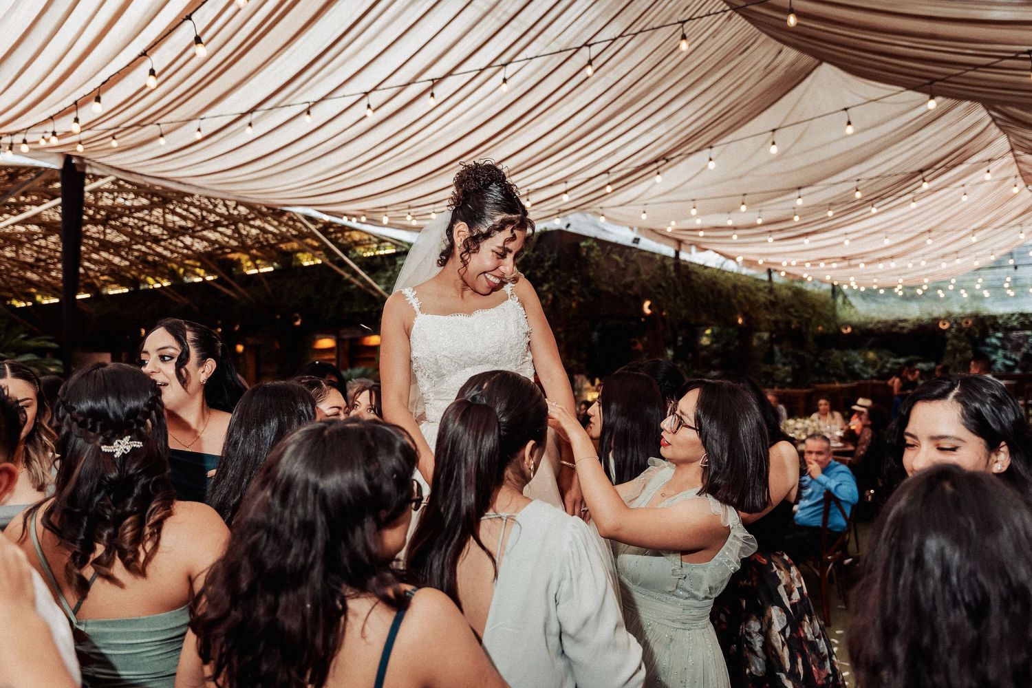 Wedding guests dancing and celebrating under draped fabric ceiling decorations and string lights at reception party.
