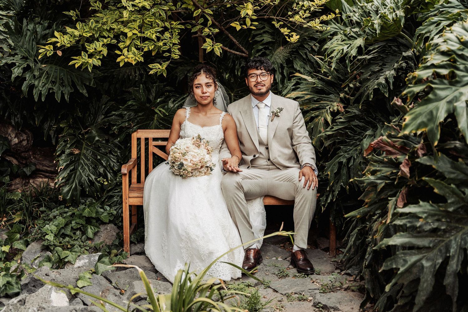 A couple in wedding attire sits on a wooden bench surrounded by lush tropical greenery and plants.
