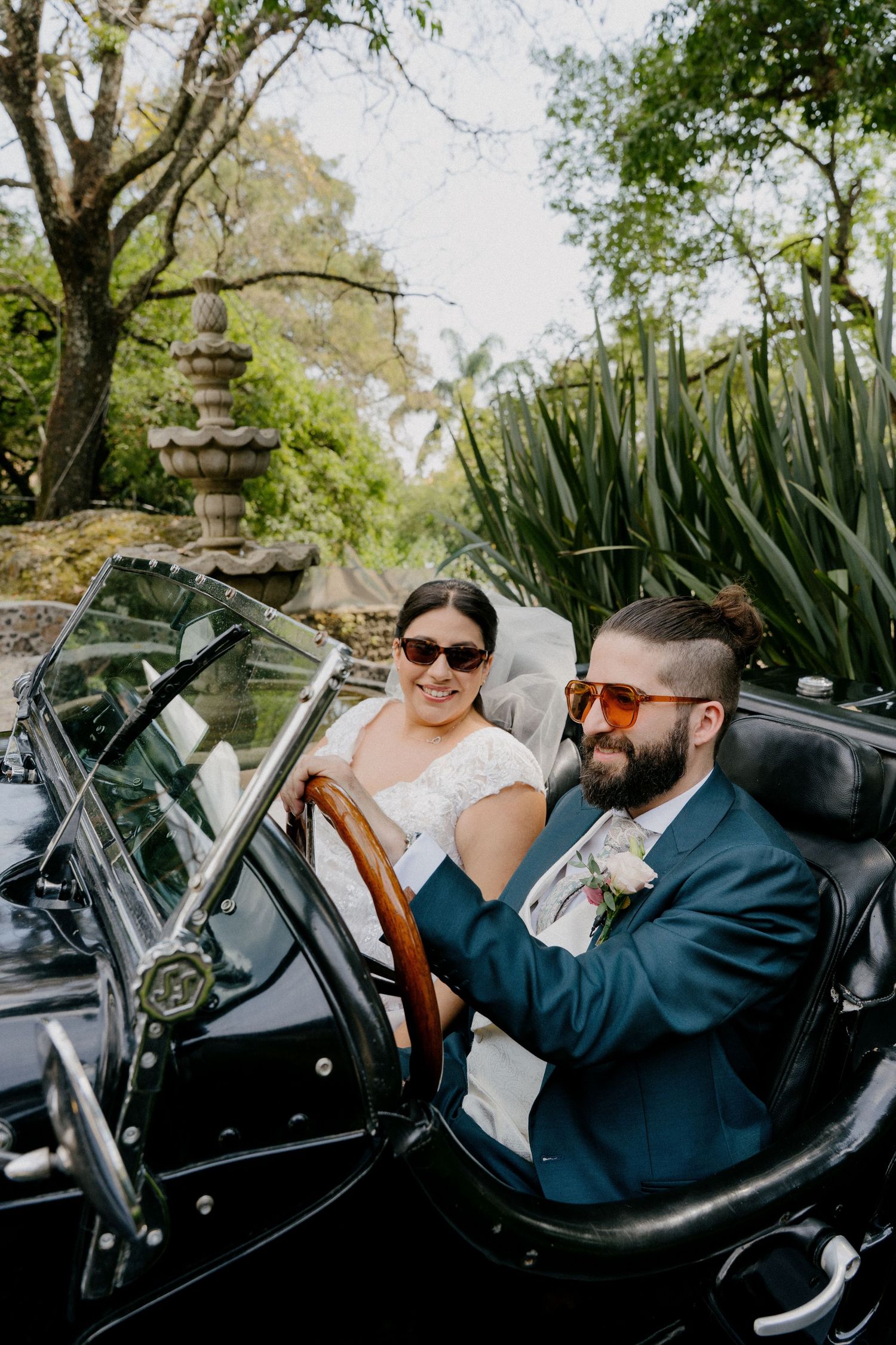 A couple sits in a vintage black convertible car surrounded by lush garden greenery and a stone pagoda.