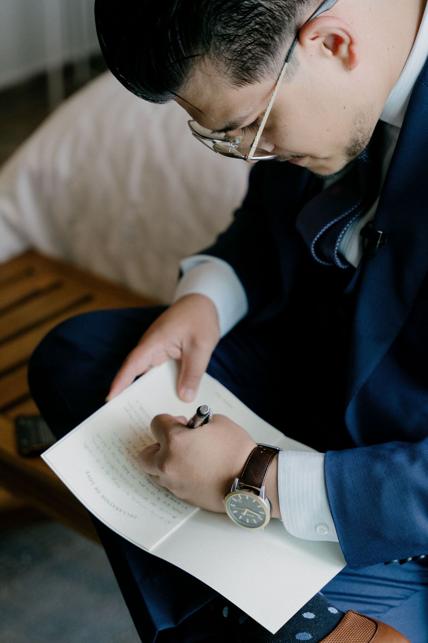 Person wearing glasses and a blue suit writes in a notebook with a wristwatch visible.