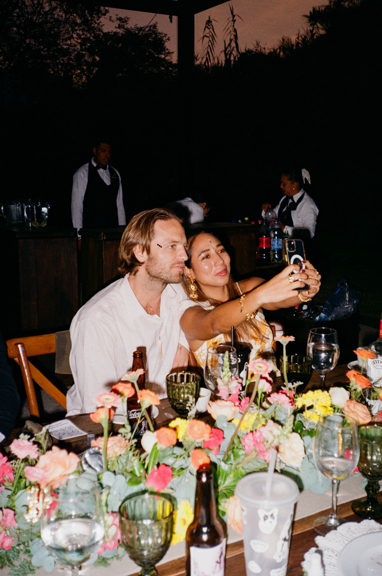 A festive dinner table decorated with colorful floral arrangements and glassware at an outdoor evening gathering.