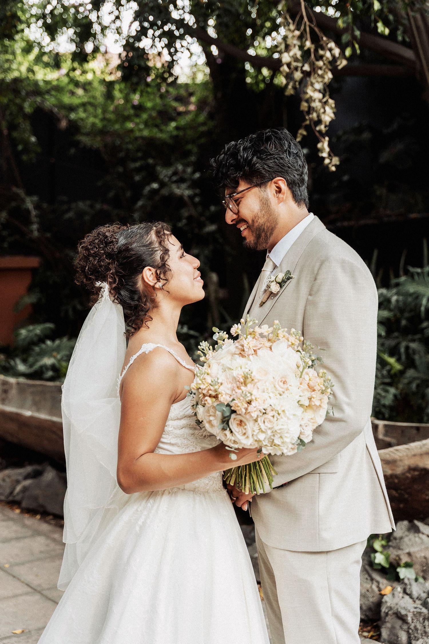 A joyful wedding moment showing a bride in white dress holding pastel bouquet with partner in light beige suit outdoors.