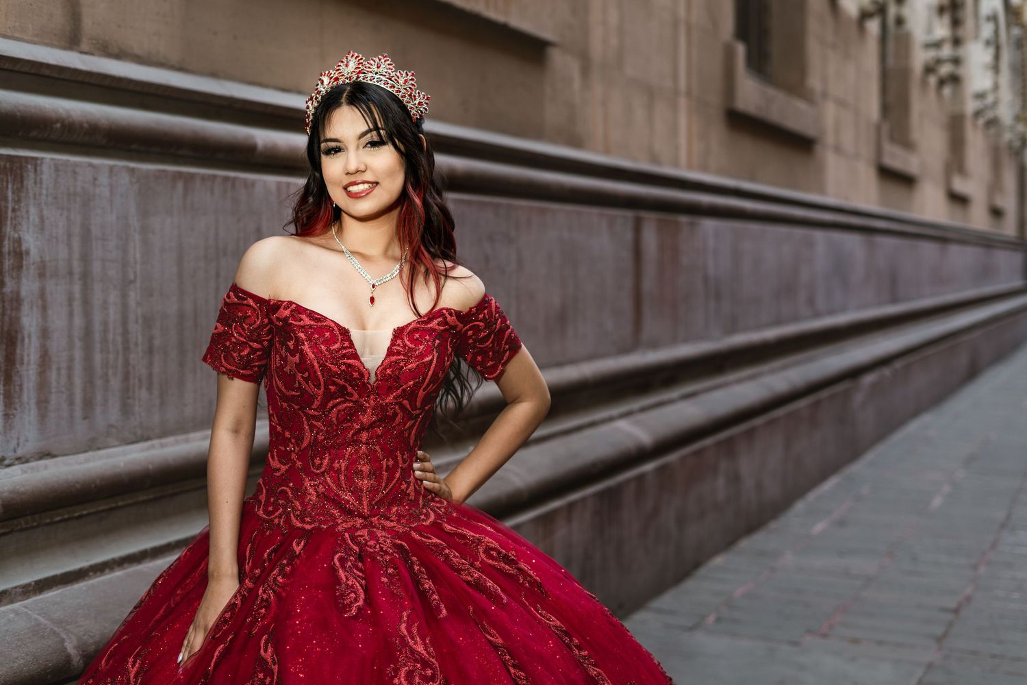 Someone poses in a luxurious off-shoulder red ball gown with intricate beading and a tiara against an urban backdrop.