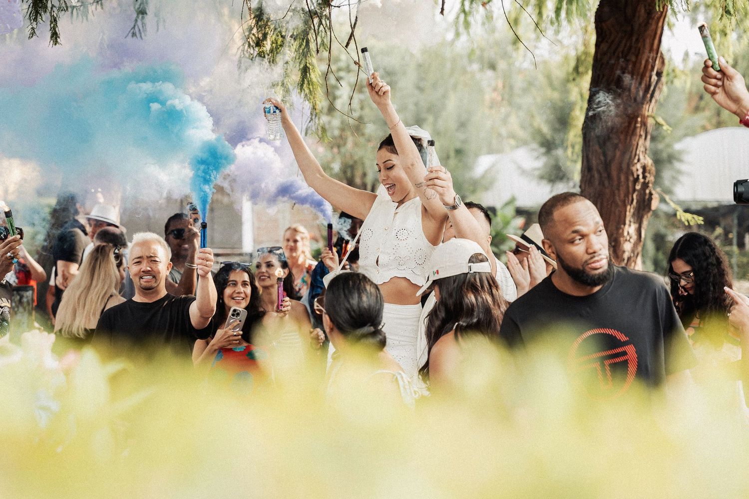 Crowd at outdoor festival with blue smoke effects and blurred yellow foreground plants.
