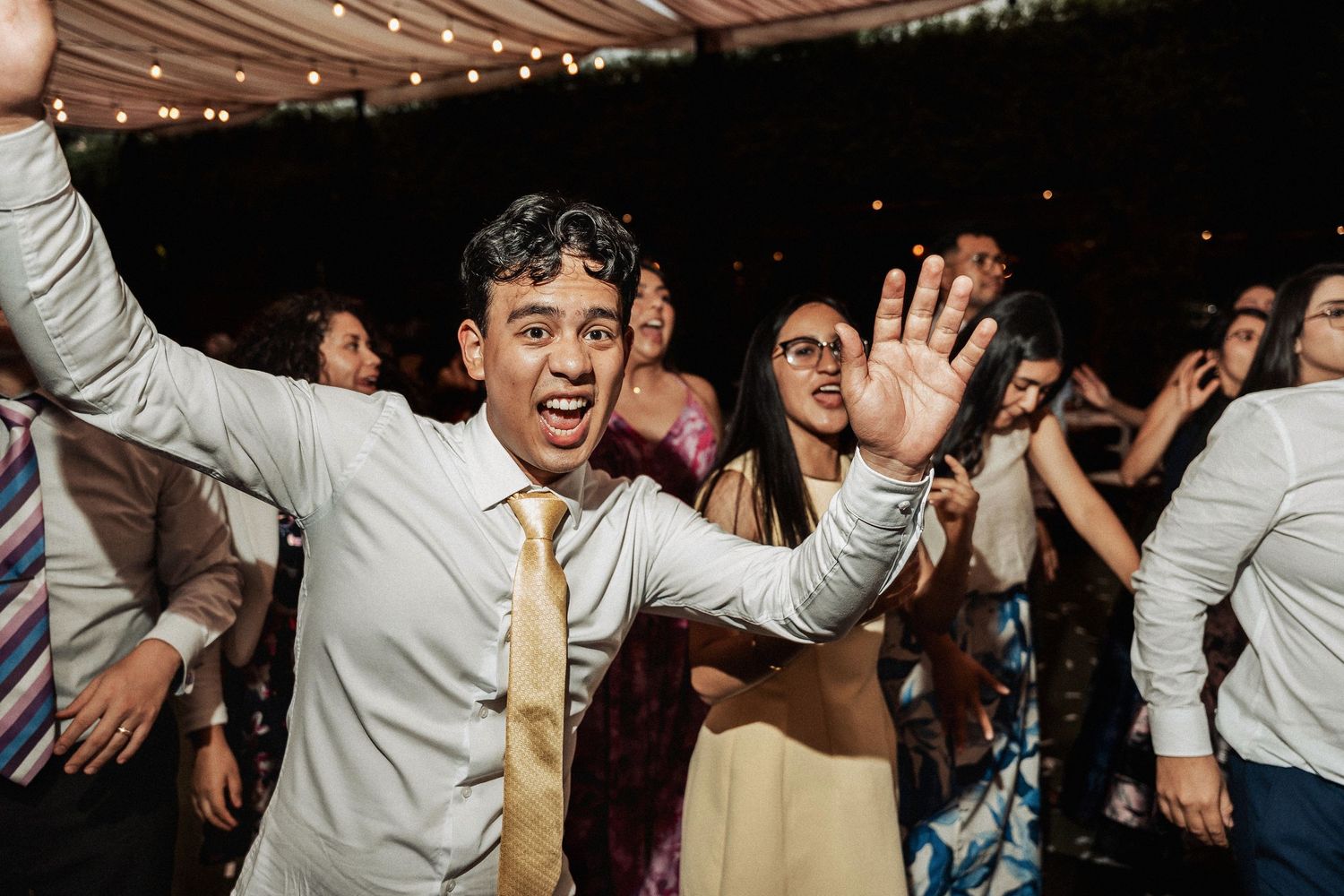 Party crowd dancing and celebrating with upbeat energy under string lights at a wedding reception.
