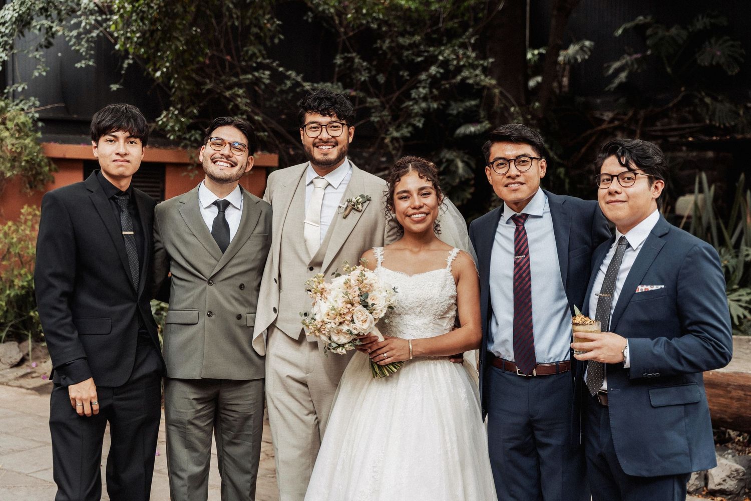 Wedding party in formal attire poses together outdoors with smiling bride holding bouquet.