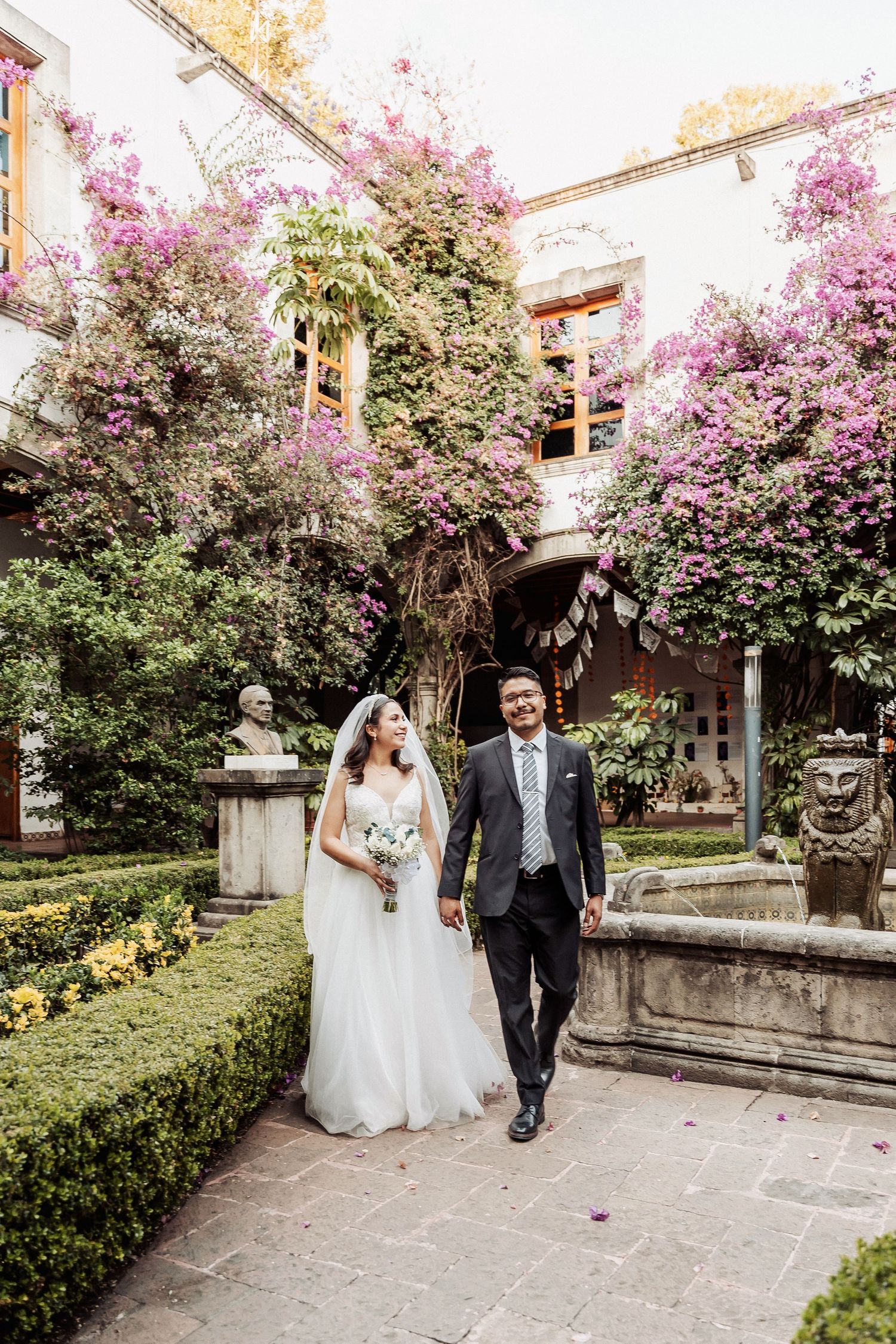 A couple walks through a beautiful courtyard garden with blooming pink flowers and manicured hedges.