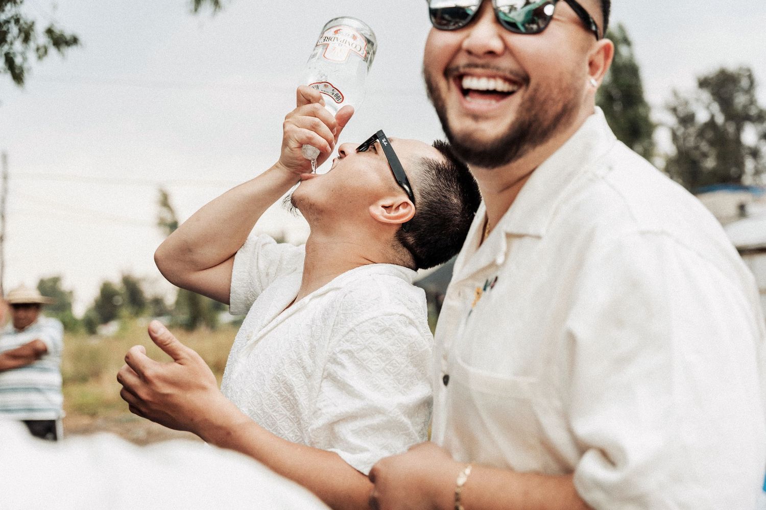 An engaged couple in white outfits share a joyful laugh while holding champagne glasses outdoors.