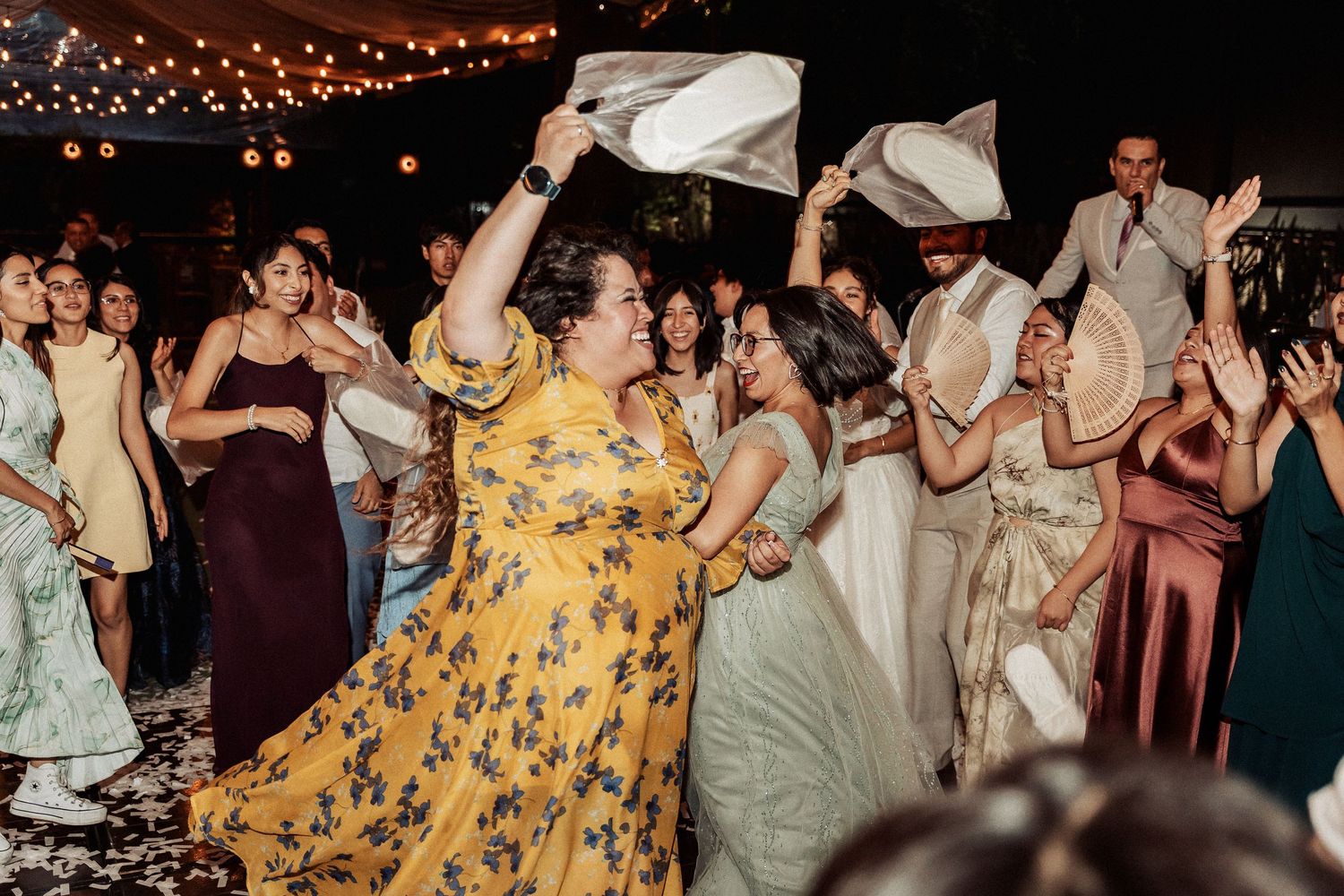 Festive wedding reception with guests dancing and waving napkins under string lights at an outdoor venue.